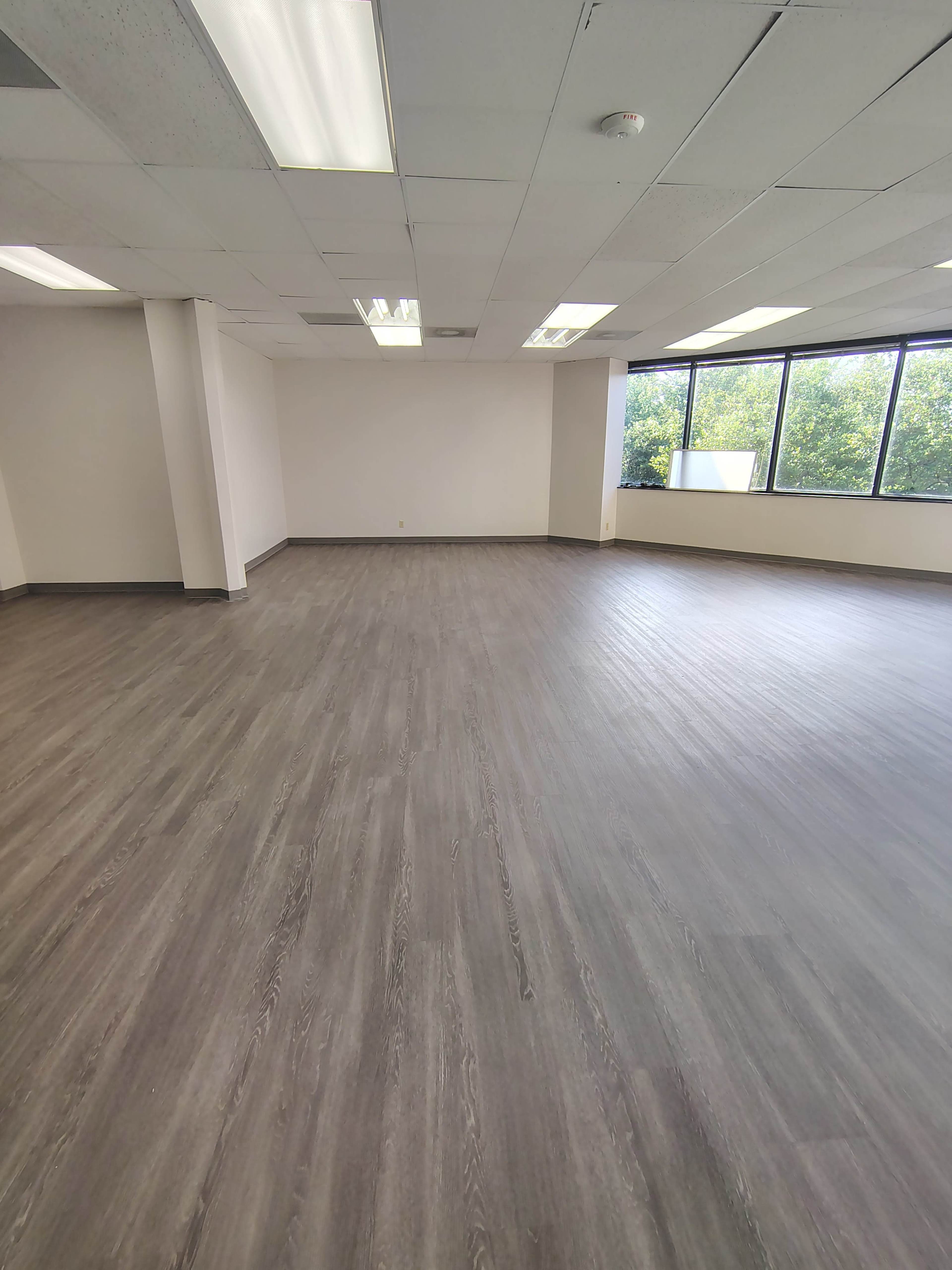 The image shows a large, empty office space with grey flooring and windows letting in natural light.