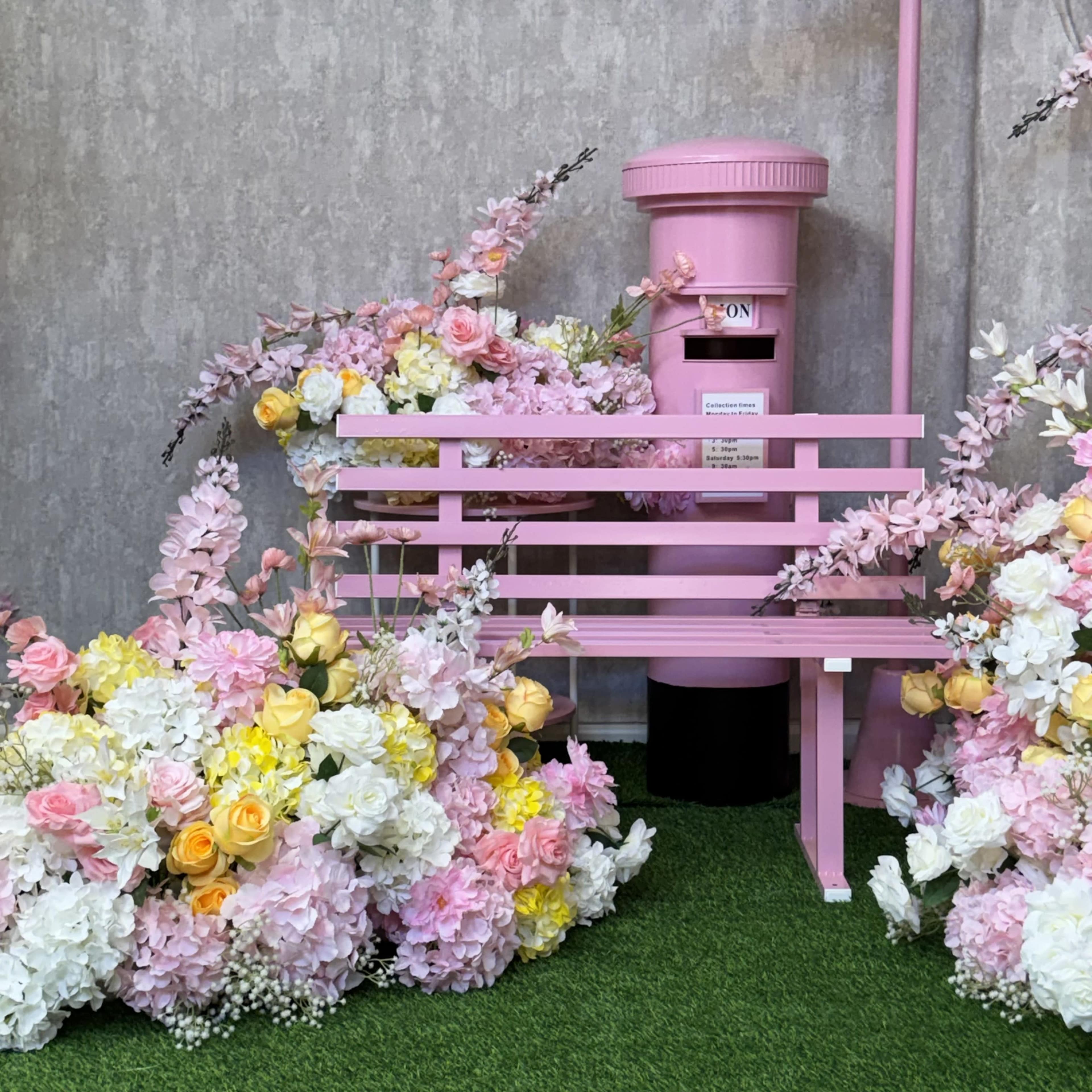The image features a pink park bench surrounded by colorful artificial flowers and a pink mail postbox.