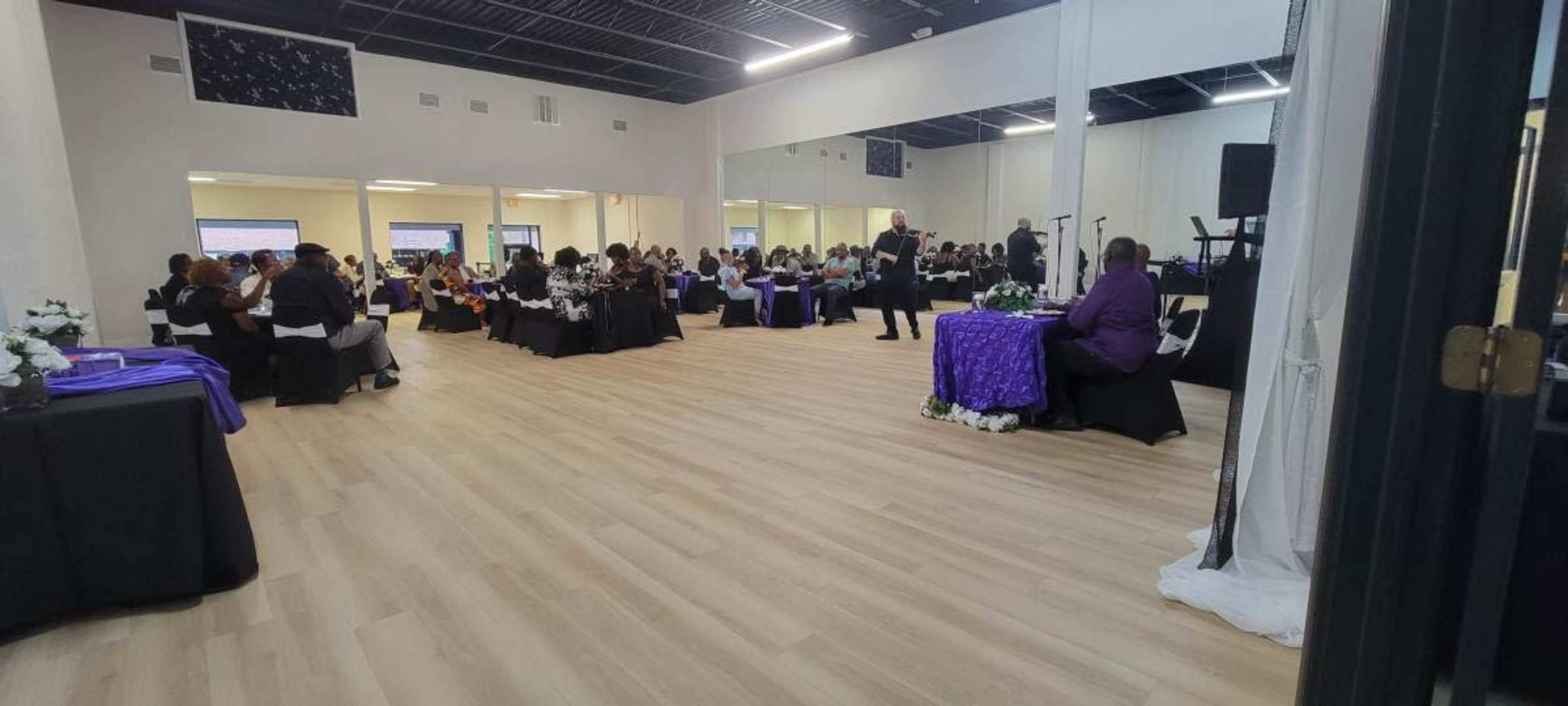 A large indoor space set up for an event with tables arranged in a dining format, featuring guests seated at various tables adorned with white and purple tablecloths.