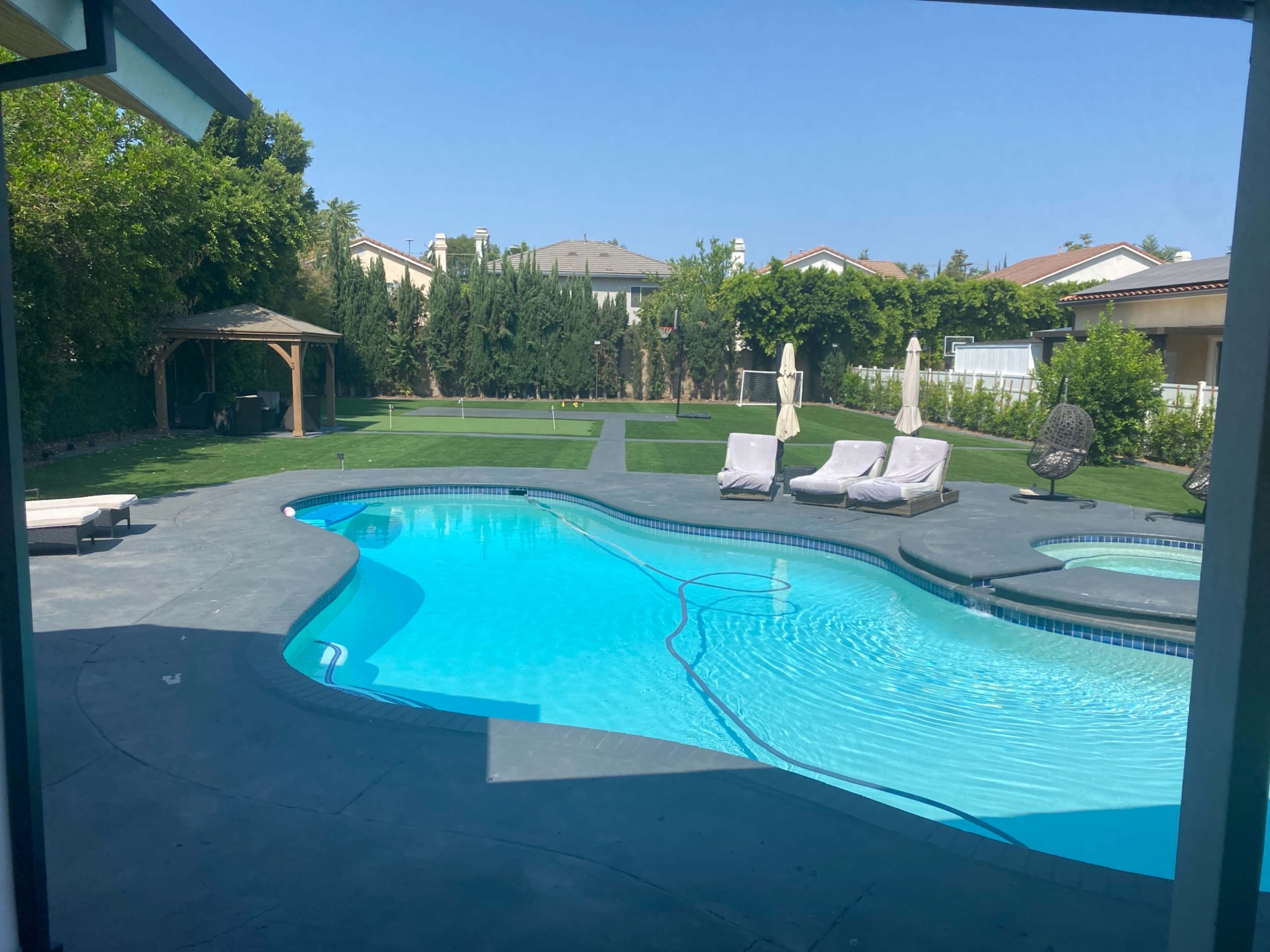 A backyard with a swimming pool surrounded by lounge chairs and grass areas, featuring a gazebo in the background.