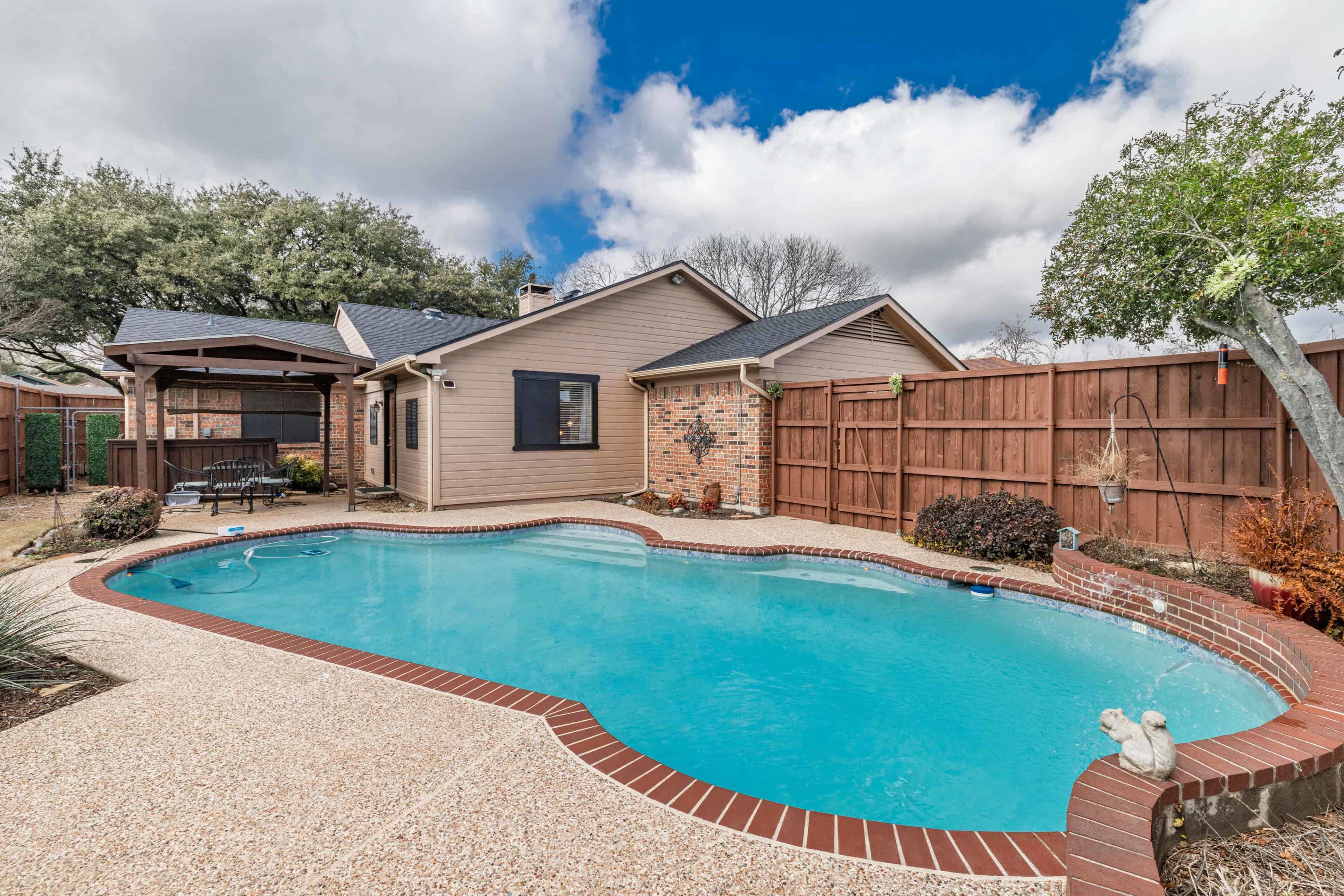 The image shows a backyard with a swimming pool surrounded by a patio, a wooden fence, and a small seating area.
