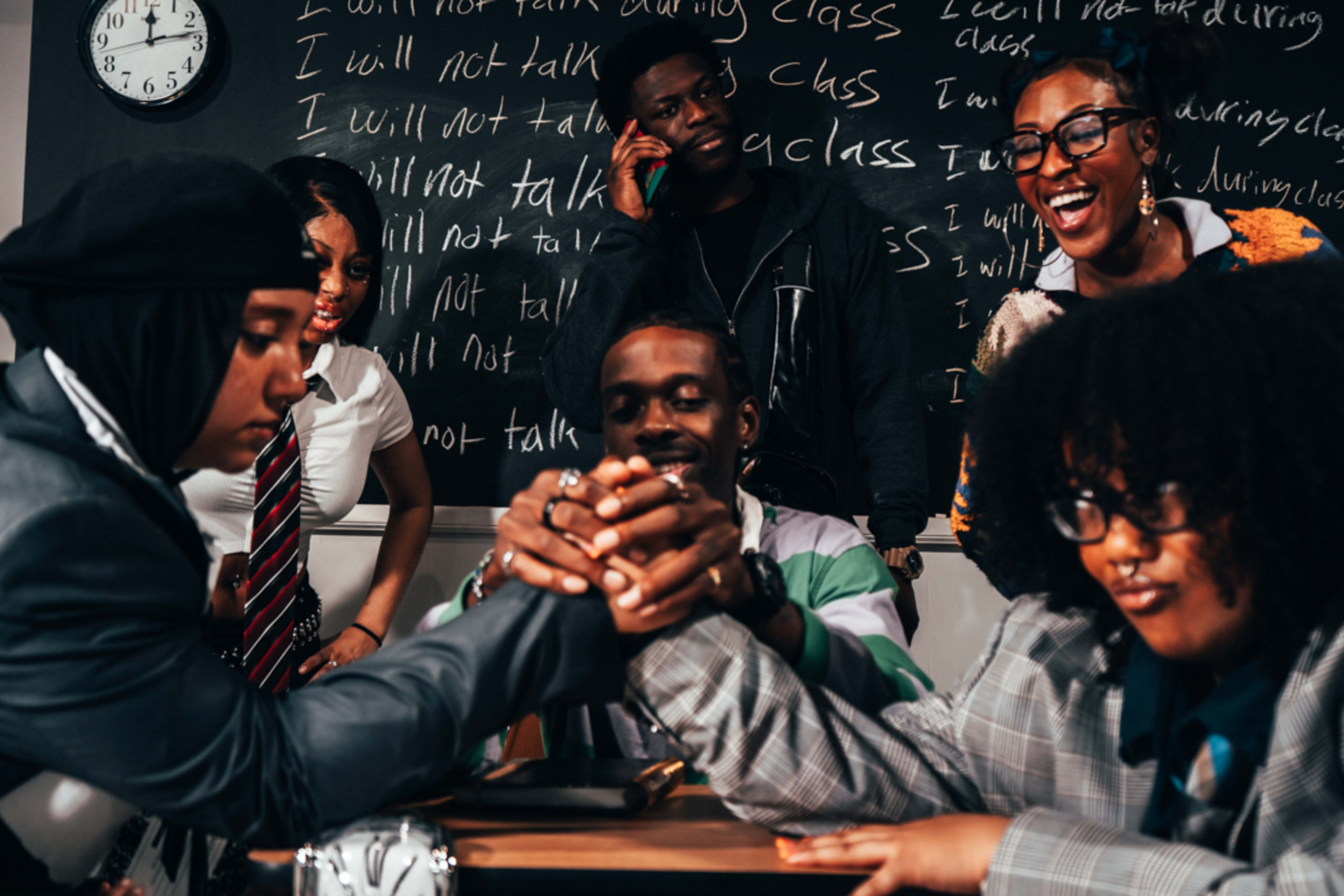 A group of six students are engaged in an arm-wrestling match at a desk in a classroom, with a blackboard filled with writing in the background.