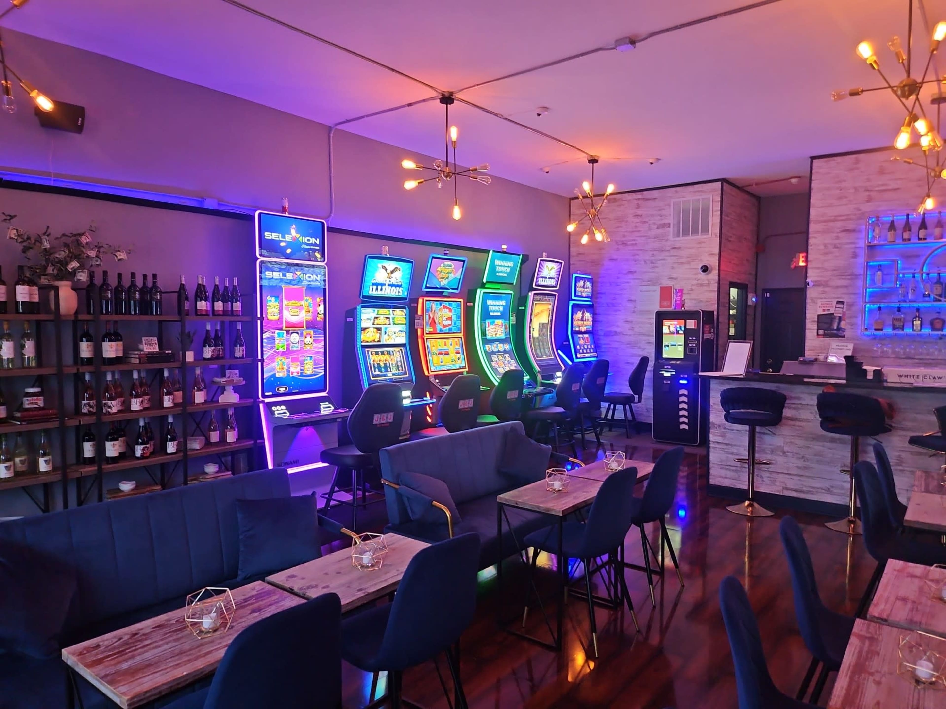 The image shows a modern venue with vibrant slot machines on one side and a seating area with tables and chairs on the other.