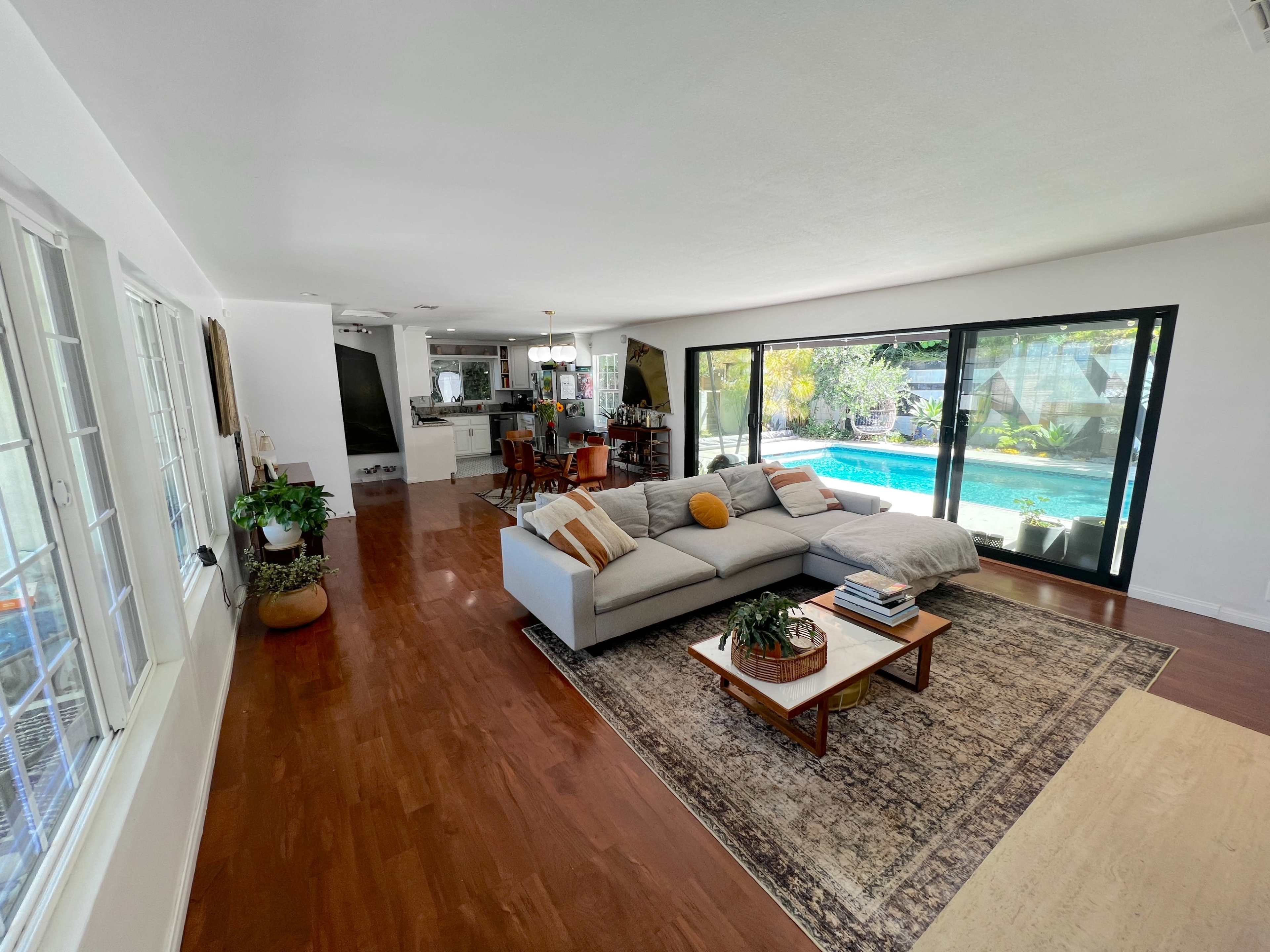 The image shows a spacious living room with a gray sofa, a coffee table, and large glass doors that open to a pool area.