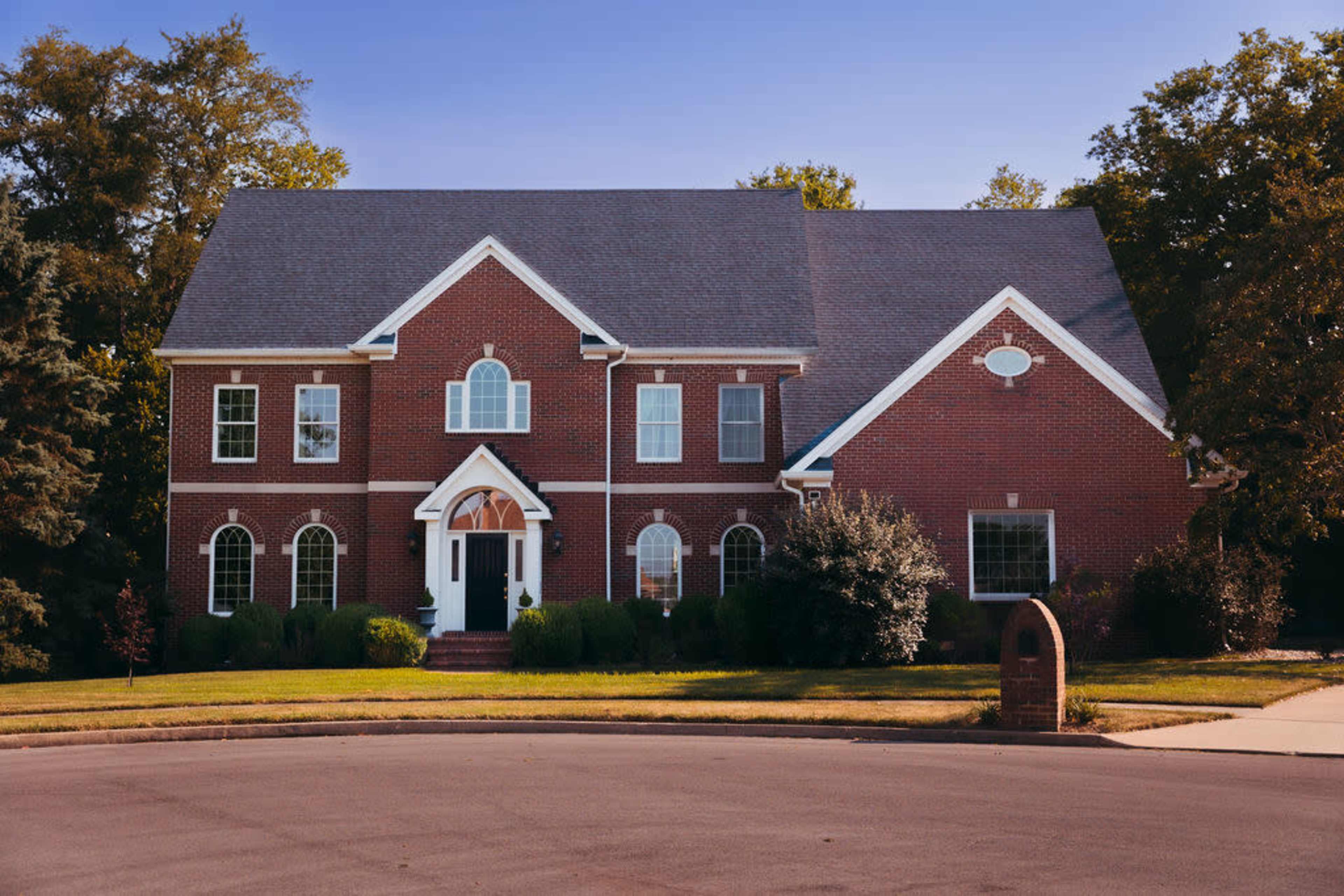 A two-story brick house with a gabled roof and multiple windows is situated on a corner lot with a landscaped front yard.