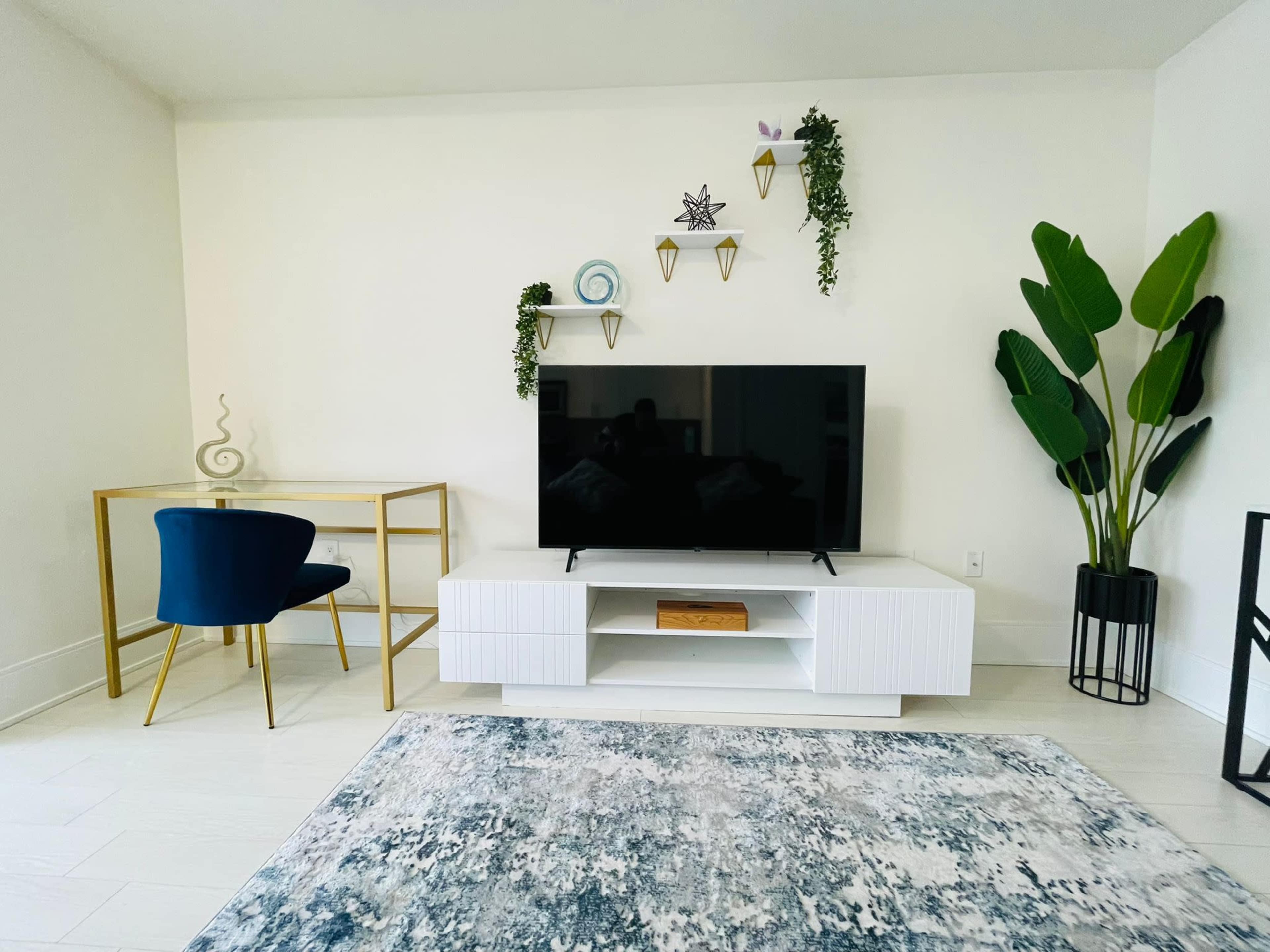 The scene features a modern living area with a white television stand, a large TV, a gold desk and chair, a patterned rug, and decorative plants on the walls and floor.