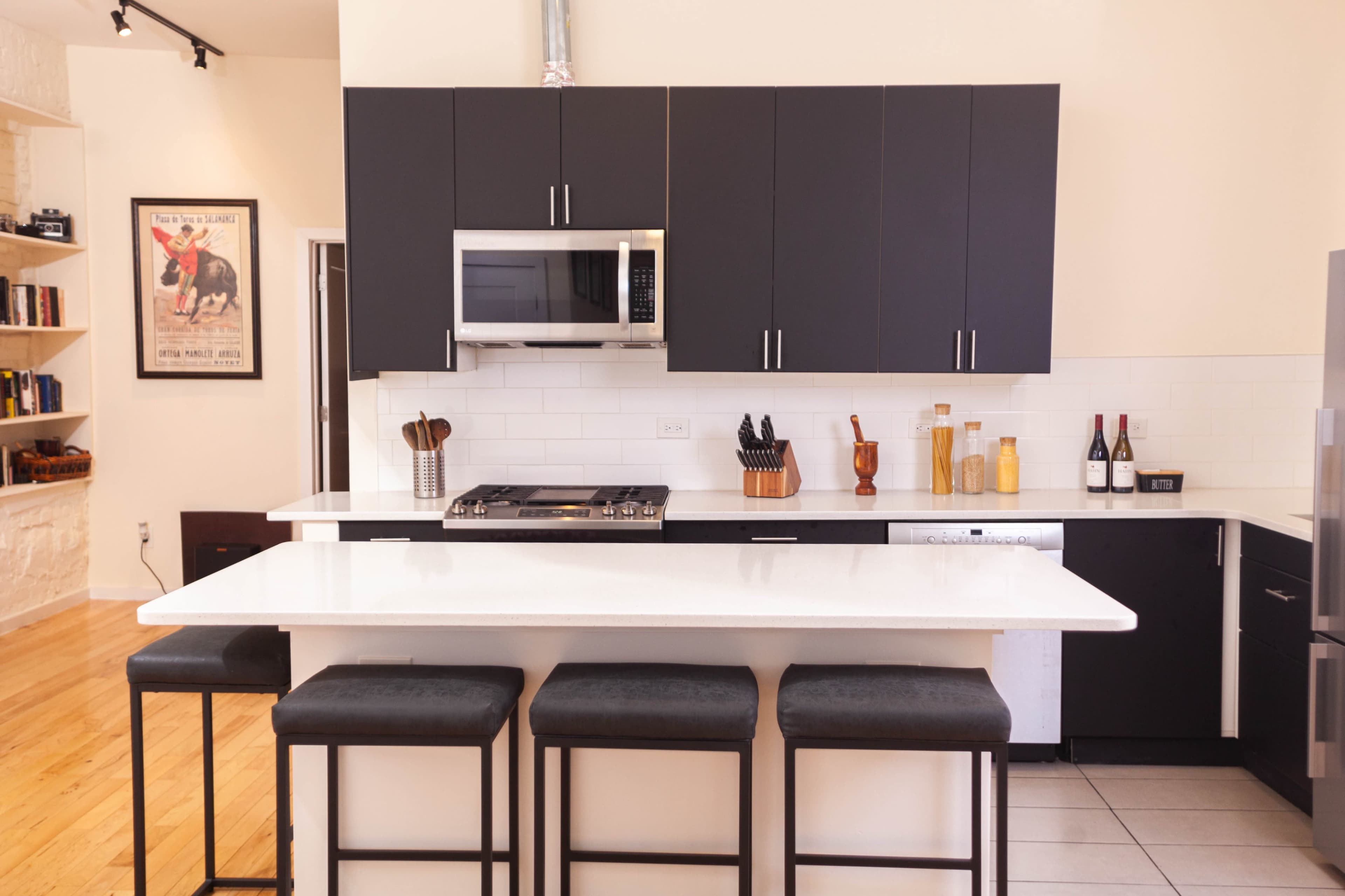 A modern kitchen with dark cabinets, a white countertop, and four black stools at the island.