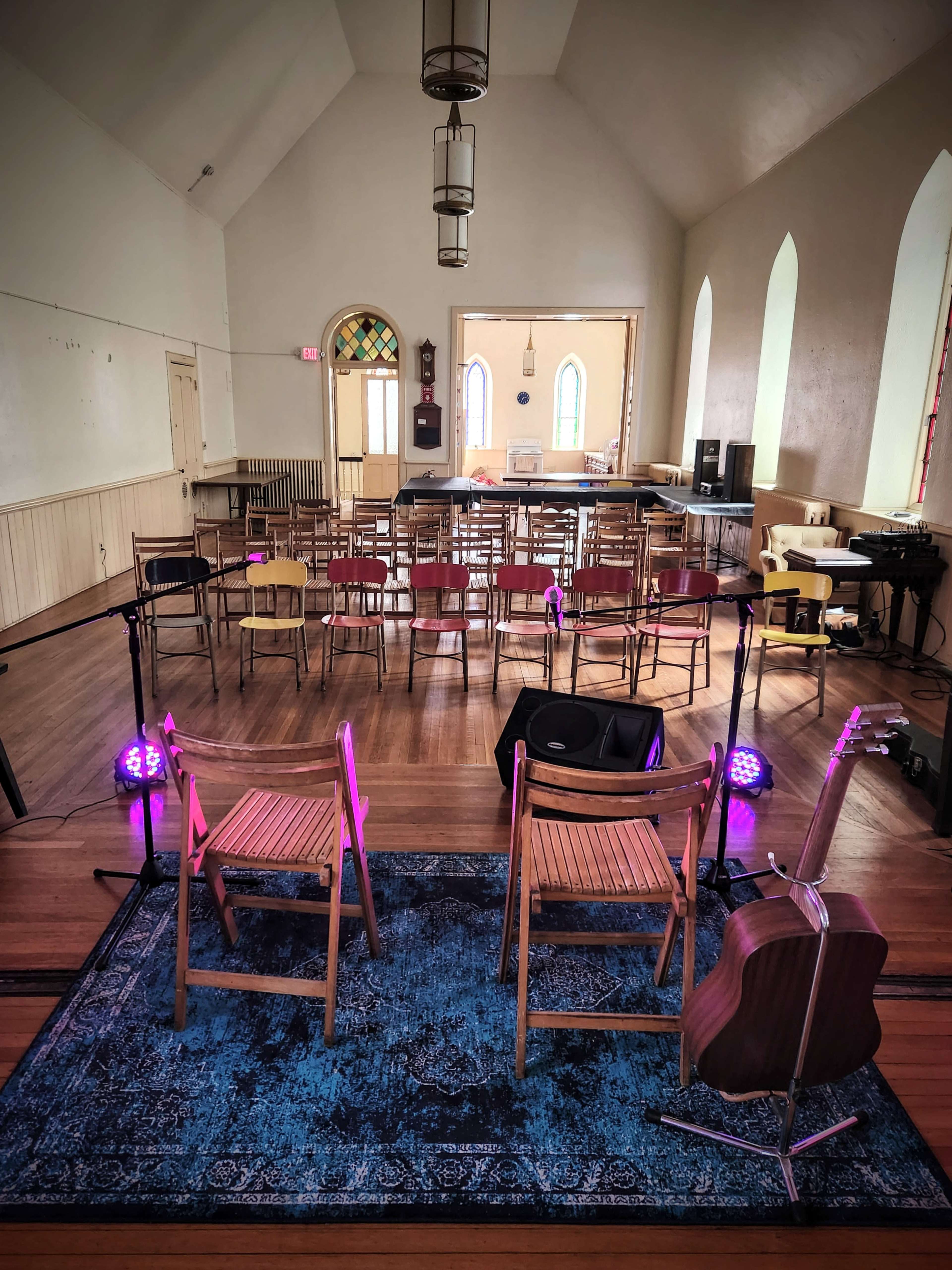 A small performance space with two wooden chairs facing an array of chairs set up for an audience, accompanied by a guitar on a stand and stage lighting.