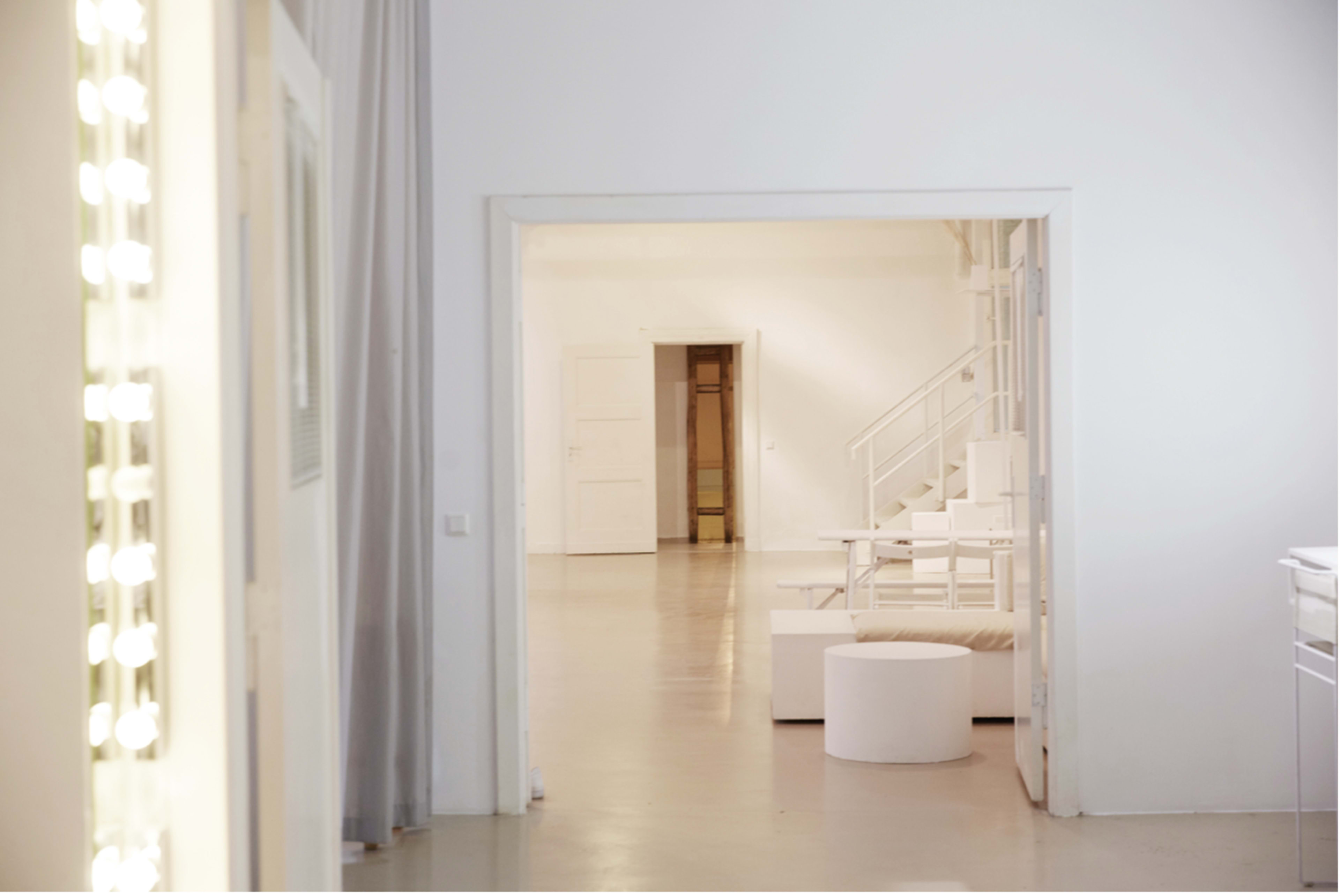 The image shows a minimalist interior space with white walls and furnishings, featuring a doorway leading to a bright area with a staircase and circular seating.