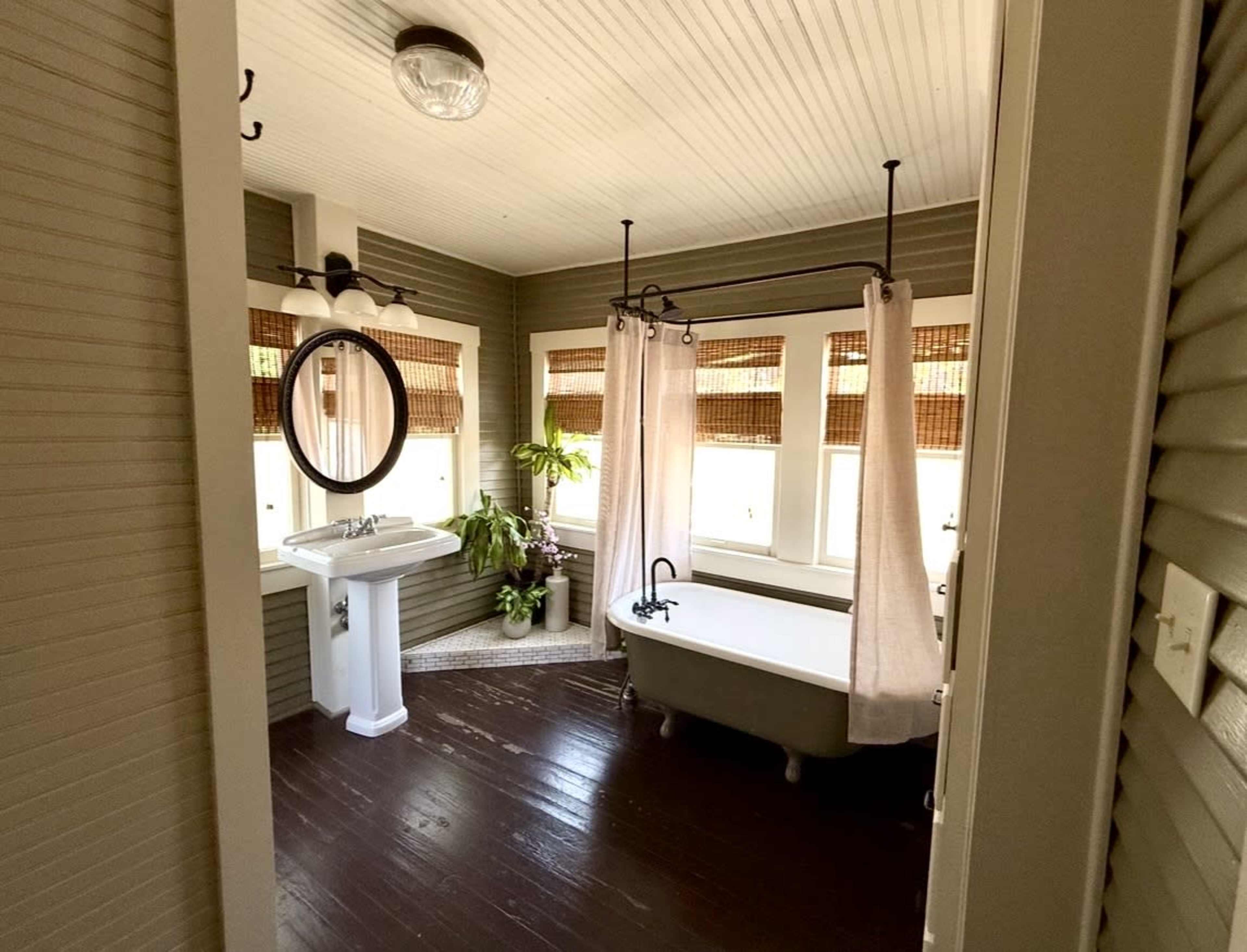 The image shows a bathroom featuring a freestanding bathtub, a pedestal sink, and large windows adorned with bamboo shades.