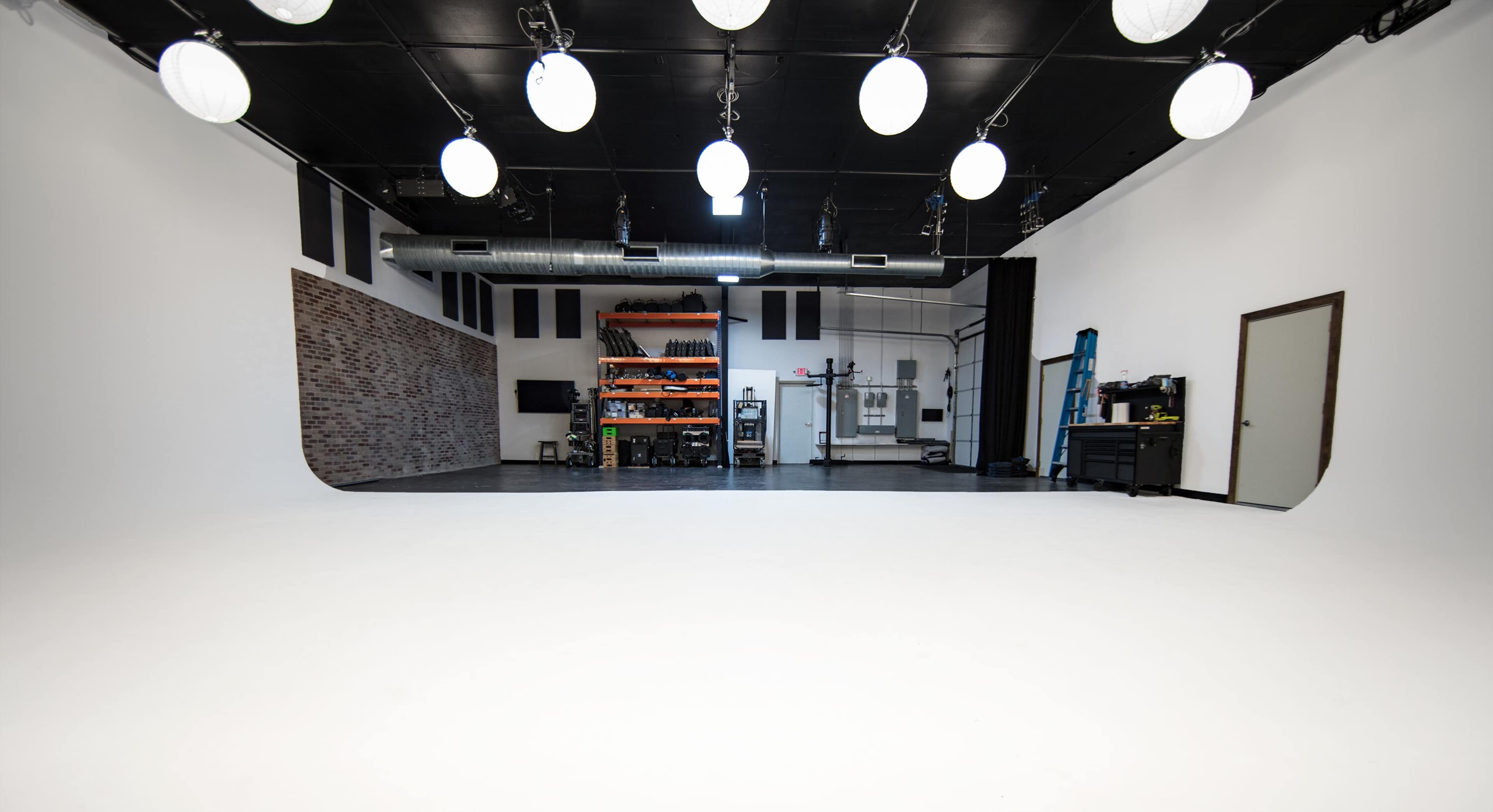 A spacious, well-lit photography studio with a white seamless backdrop, overhead lighting, and various equipment organized on a shelf.
