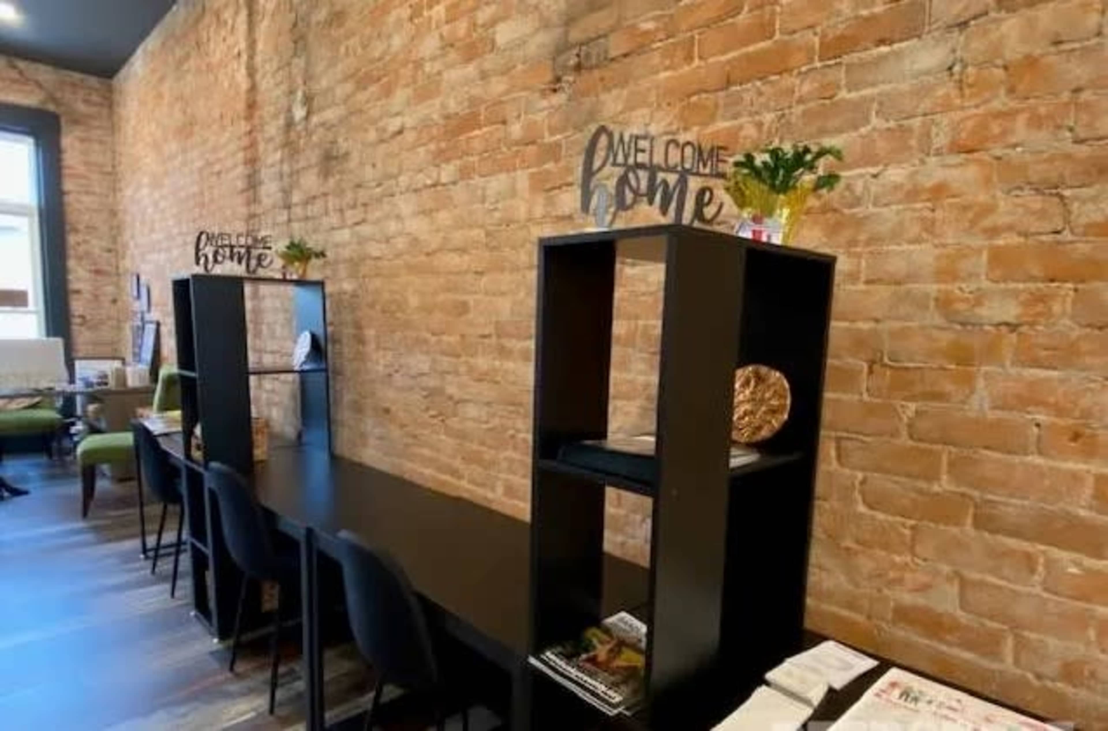 A cozy interior space with a brick wall, featuring black shelving units and a long table with chairs, adorned with decorative items and plants.