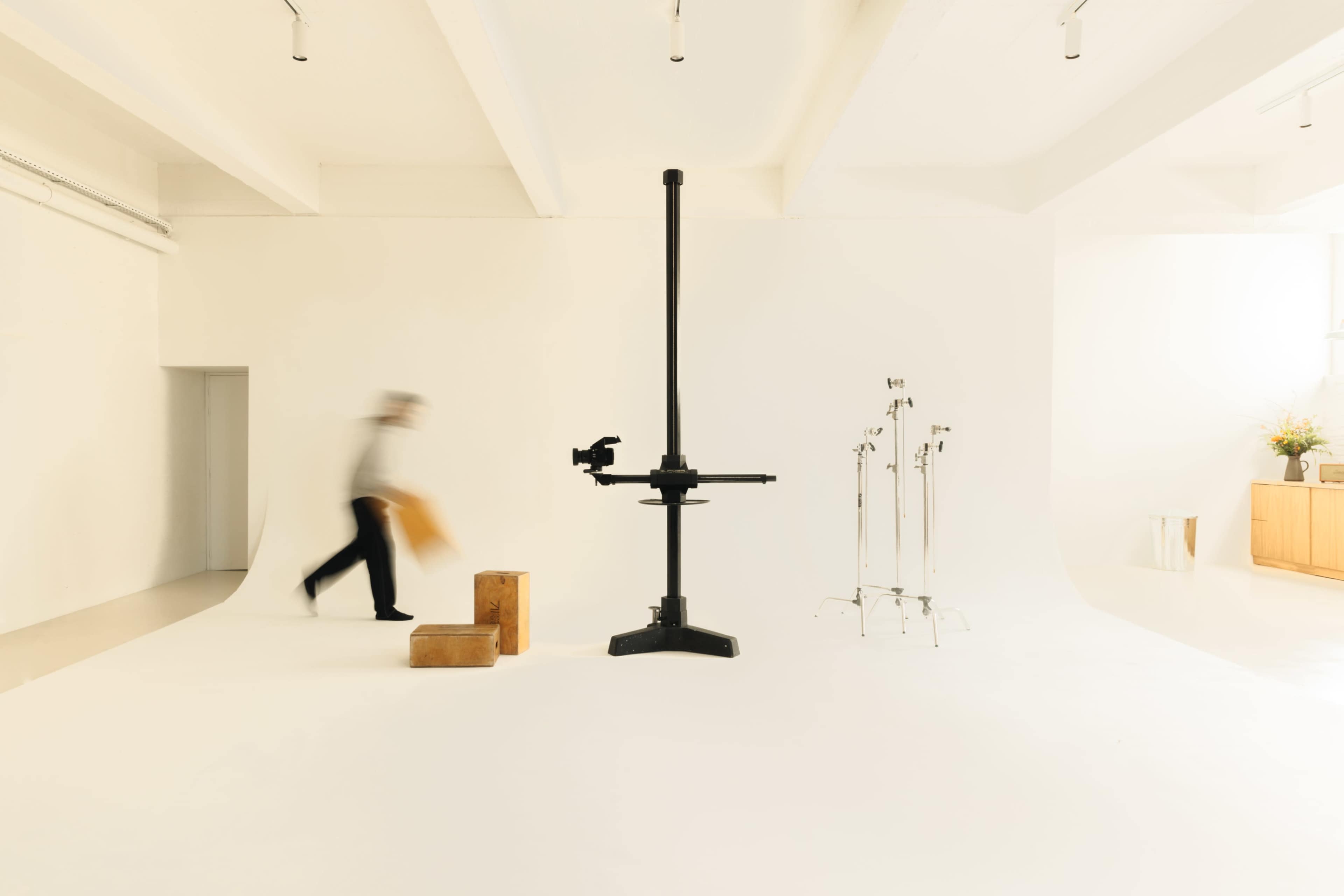 A person carrying an item walks past a large camera stand in a minimalist studio space.