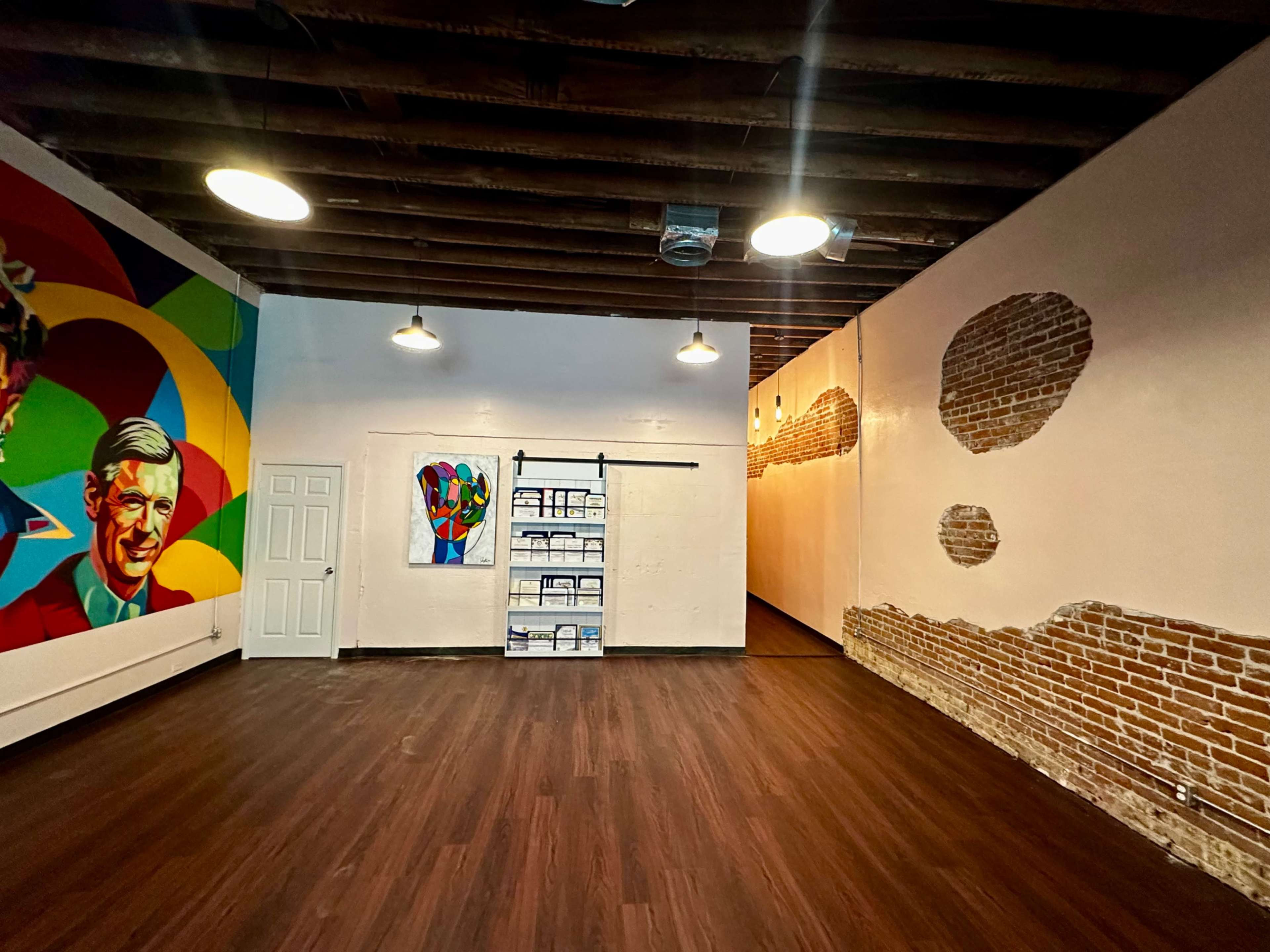The image shows a spacious room with wooden floors, featuring colorful murals on one wall and exposed brick on another.