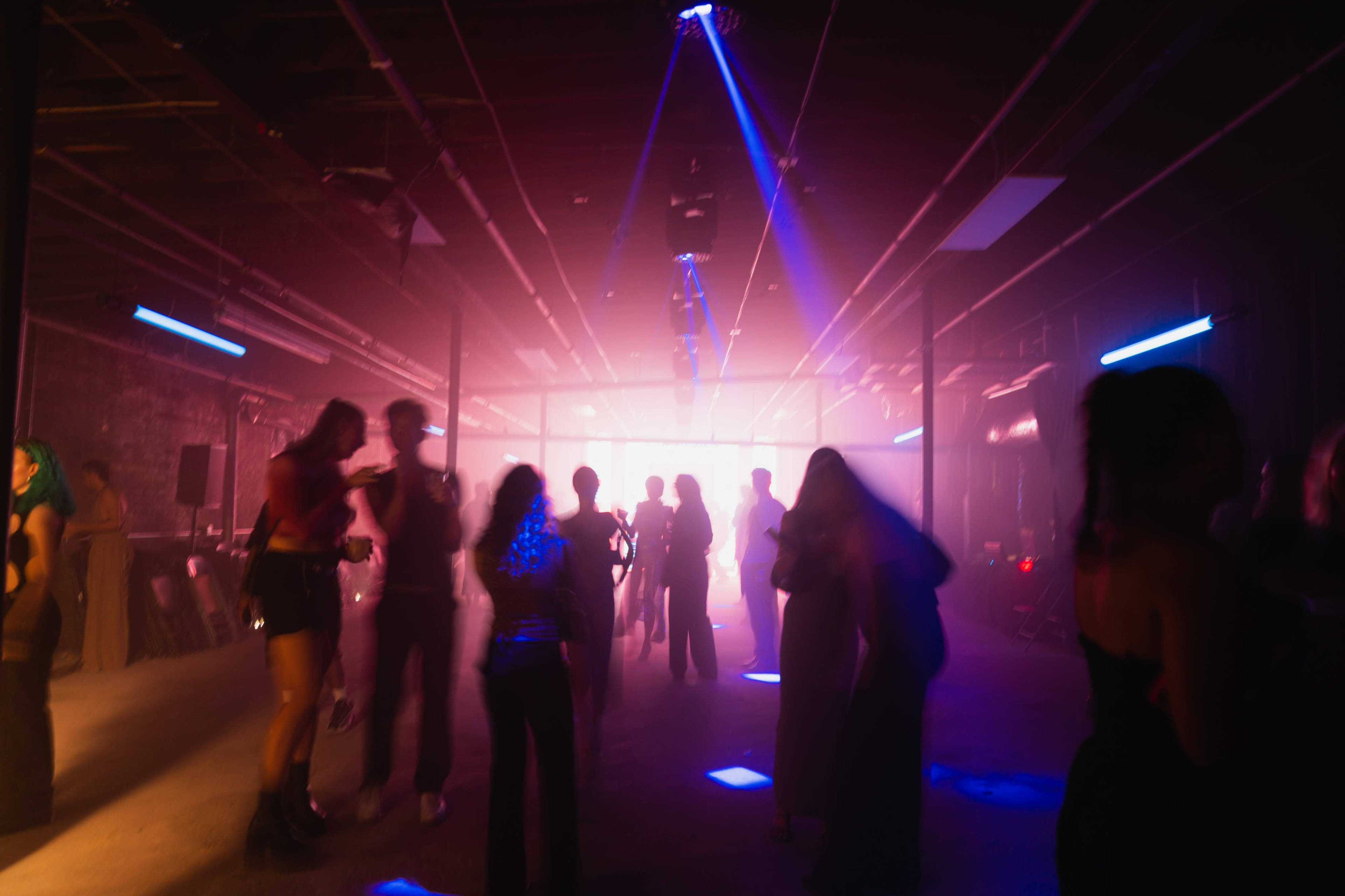 A dimly lit warehouse is filled with silhouettes of people dancing under colorful lights and a haze of fog.