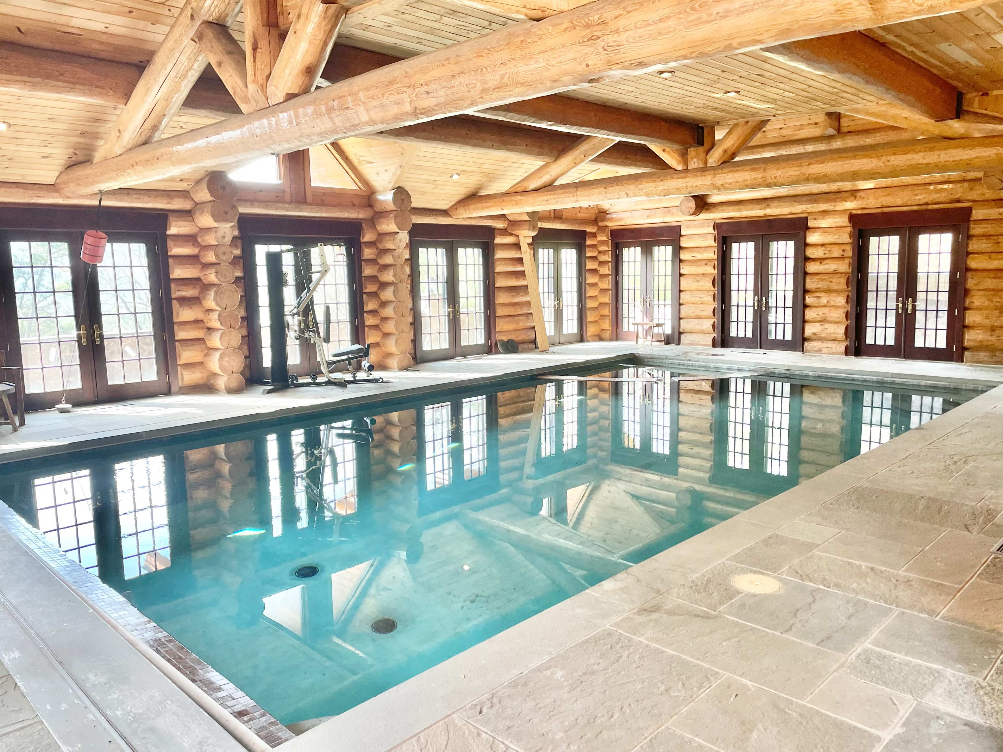 An indoor swimming pool surrounded by wooden log walls and large glass doors.