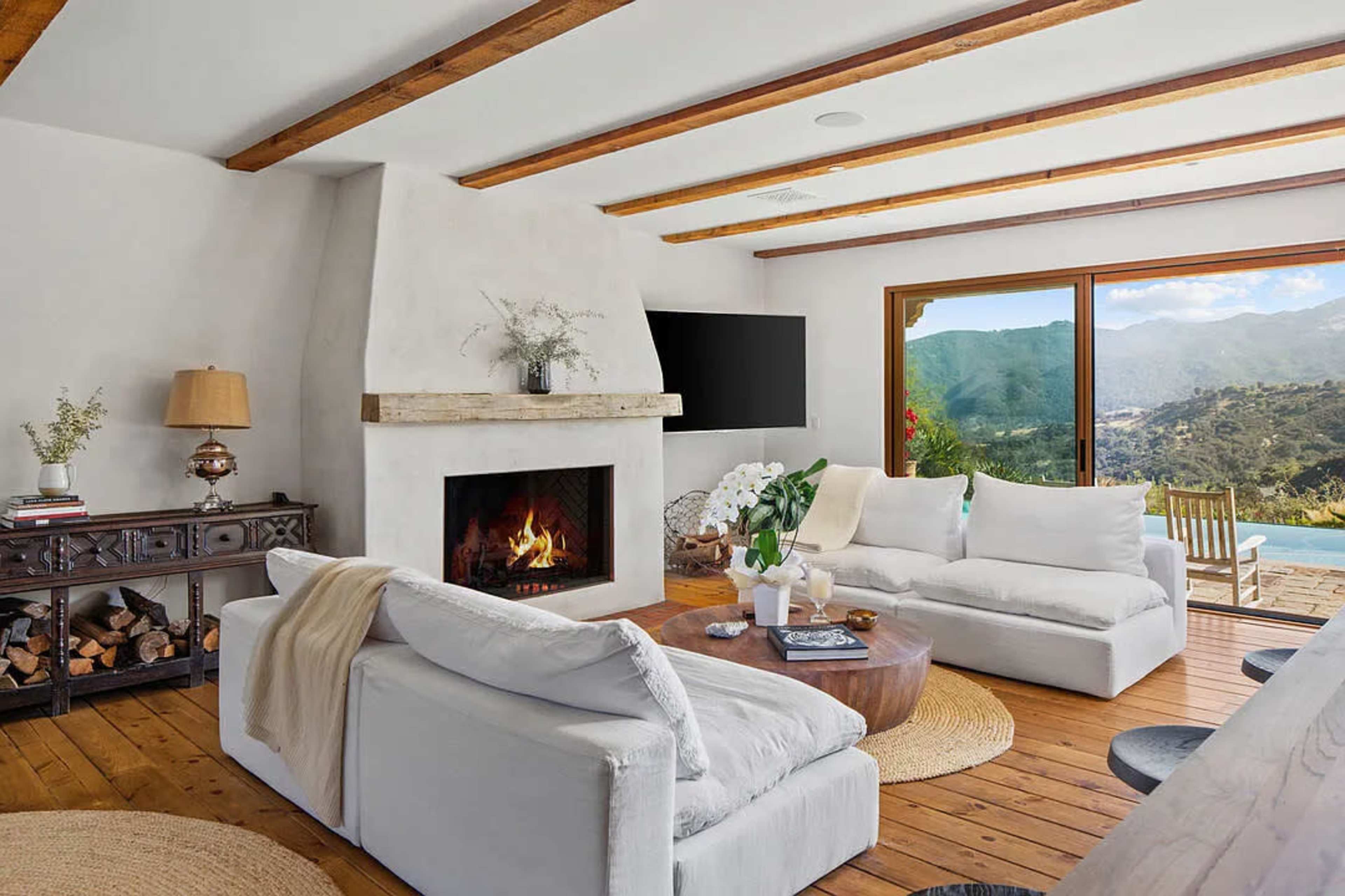 A cozy living room features a fireplace, comfortable seating, wooden ceiling beams, and a large window overlooking a mountainous view.