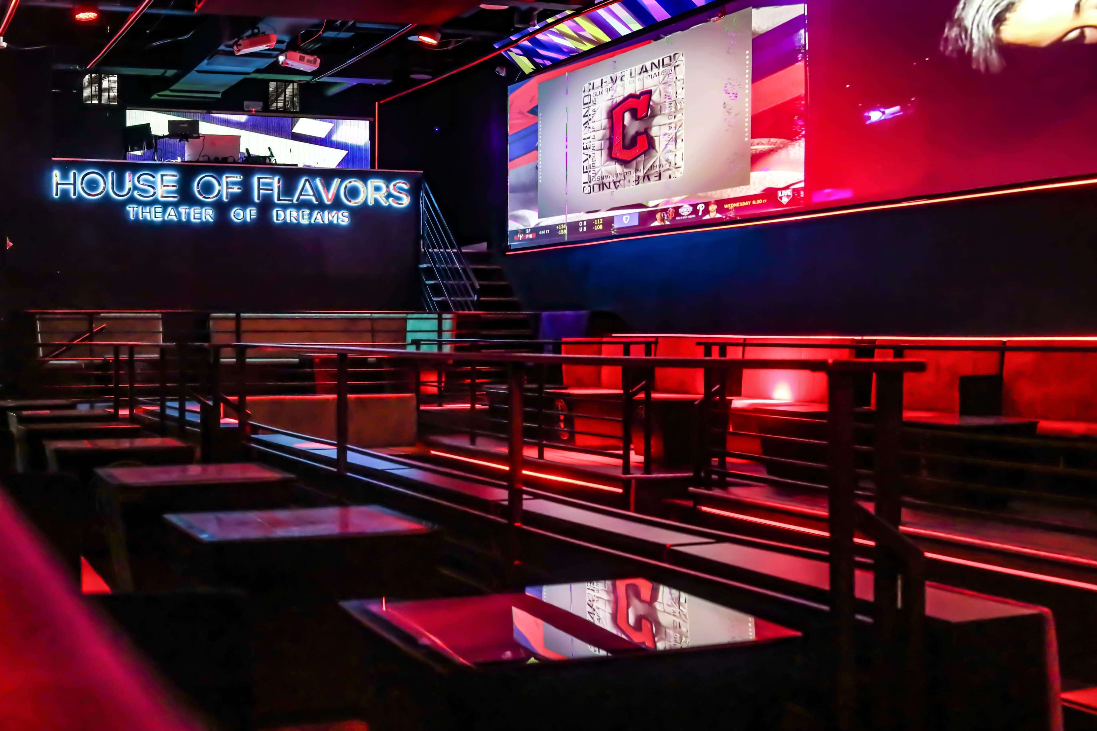 The image shows an empty interior of a modern venue called "House of Flavors" with a large screen displaying a sports logo and illuminated seating areas in red and blue lighting.