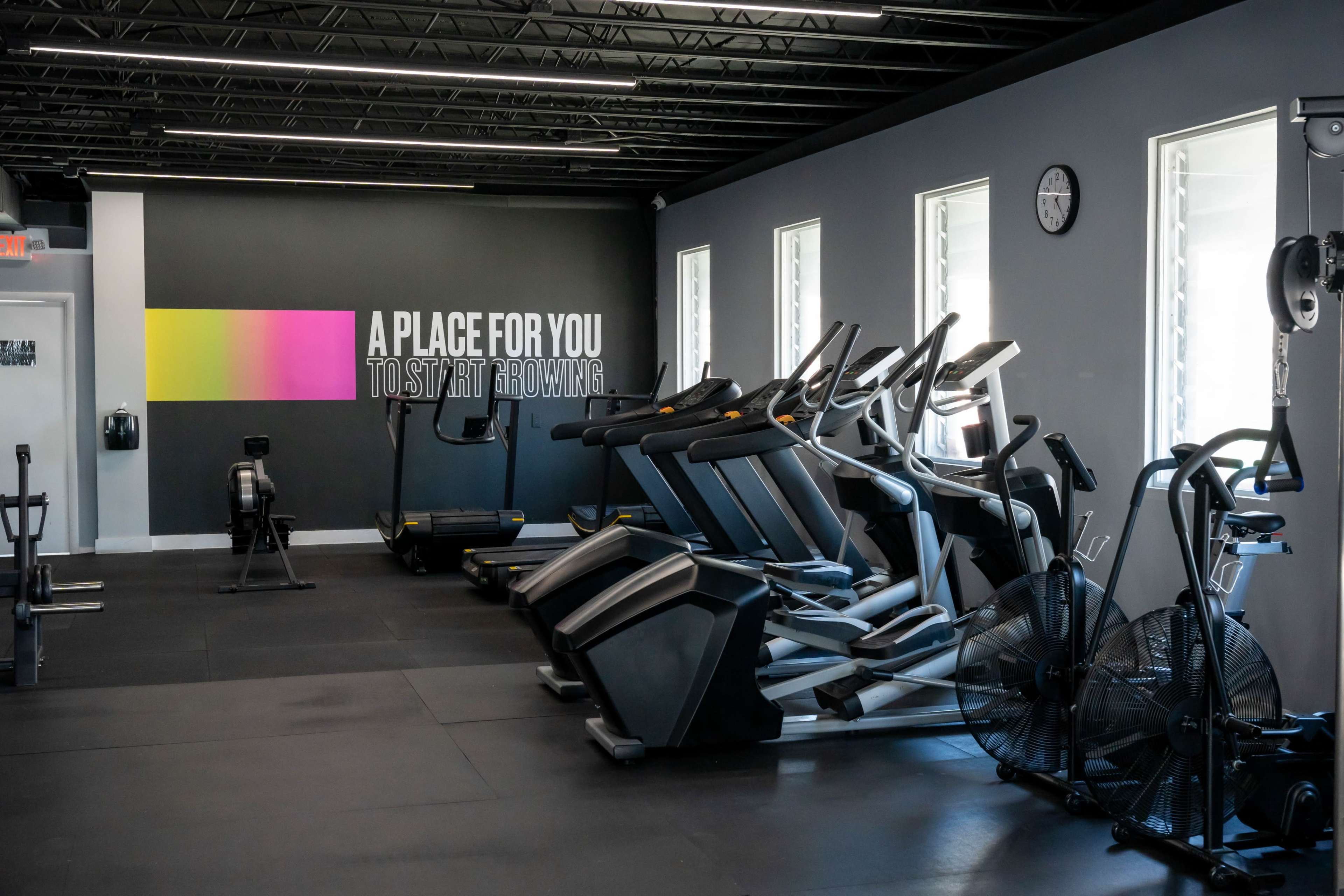 The image shows a modern gym interior featuring various exercise equipment, including treadmills and stationary bikes, against a backdrop of motivational text on the wall.
