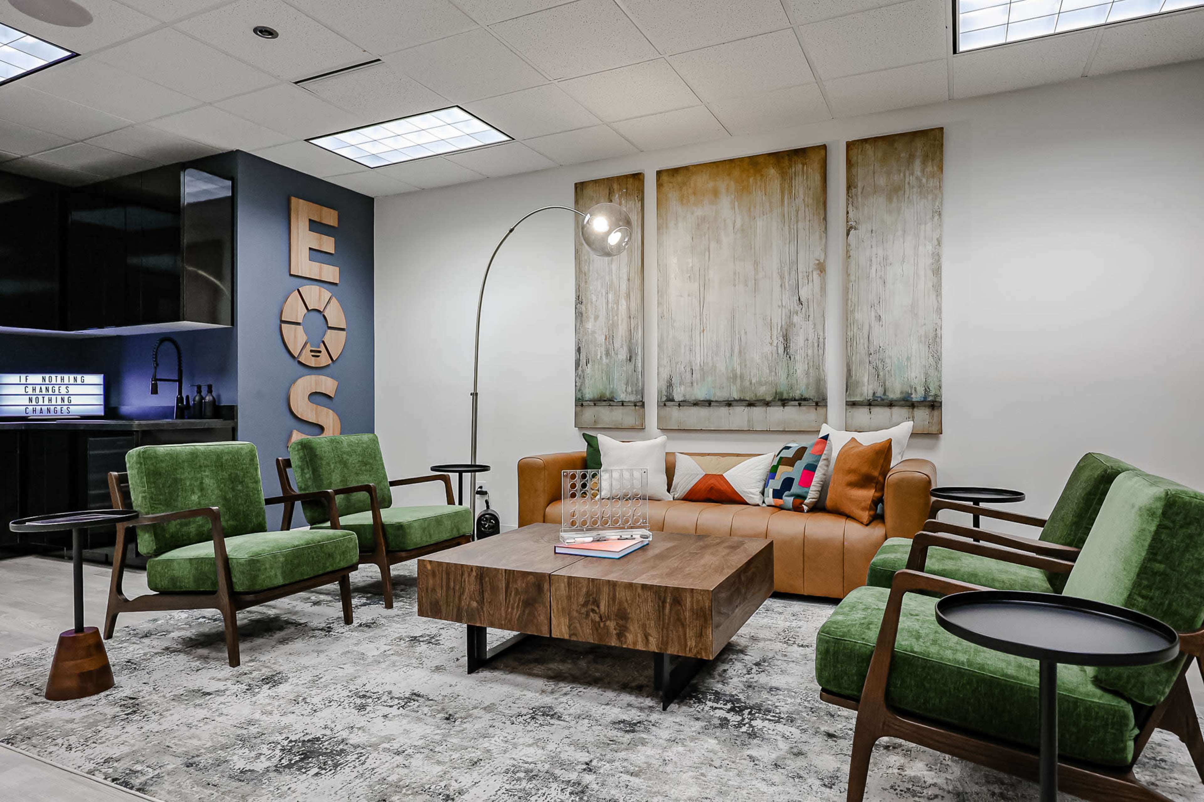 The image shows a modern lounge area with a brown leather sofa, green chairs, a wooden coffee table, and large abstract paintings on the wall.