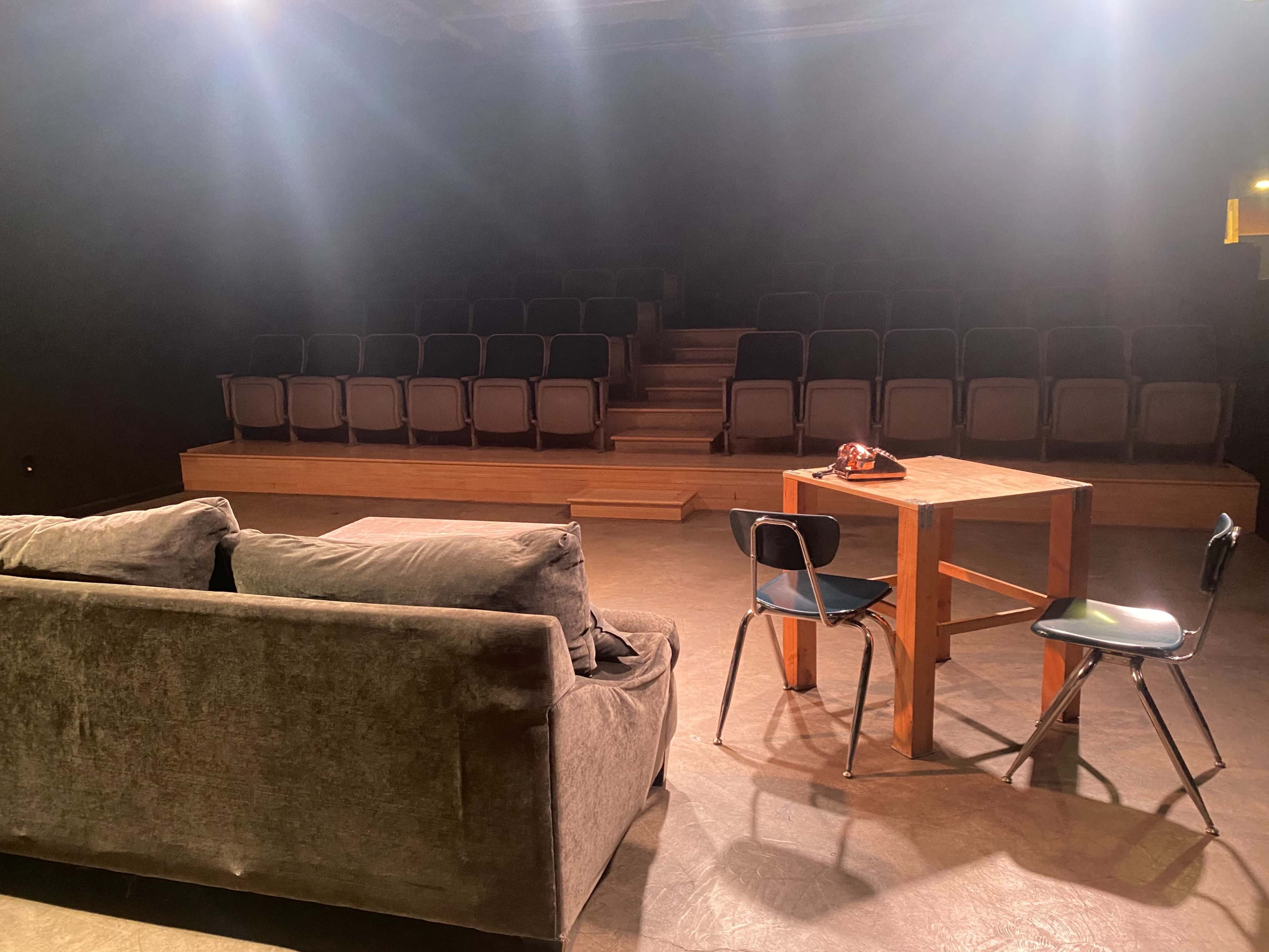 A dimly lit theater setup features a gray couch and a wooden table with chairs facing a tiered seating area.