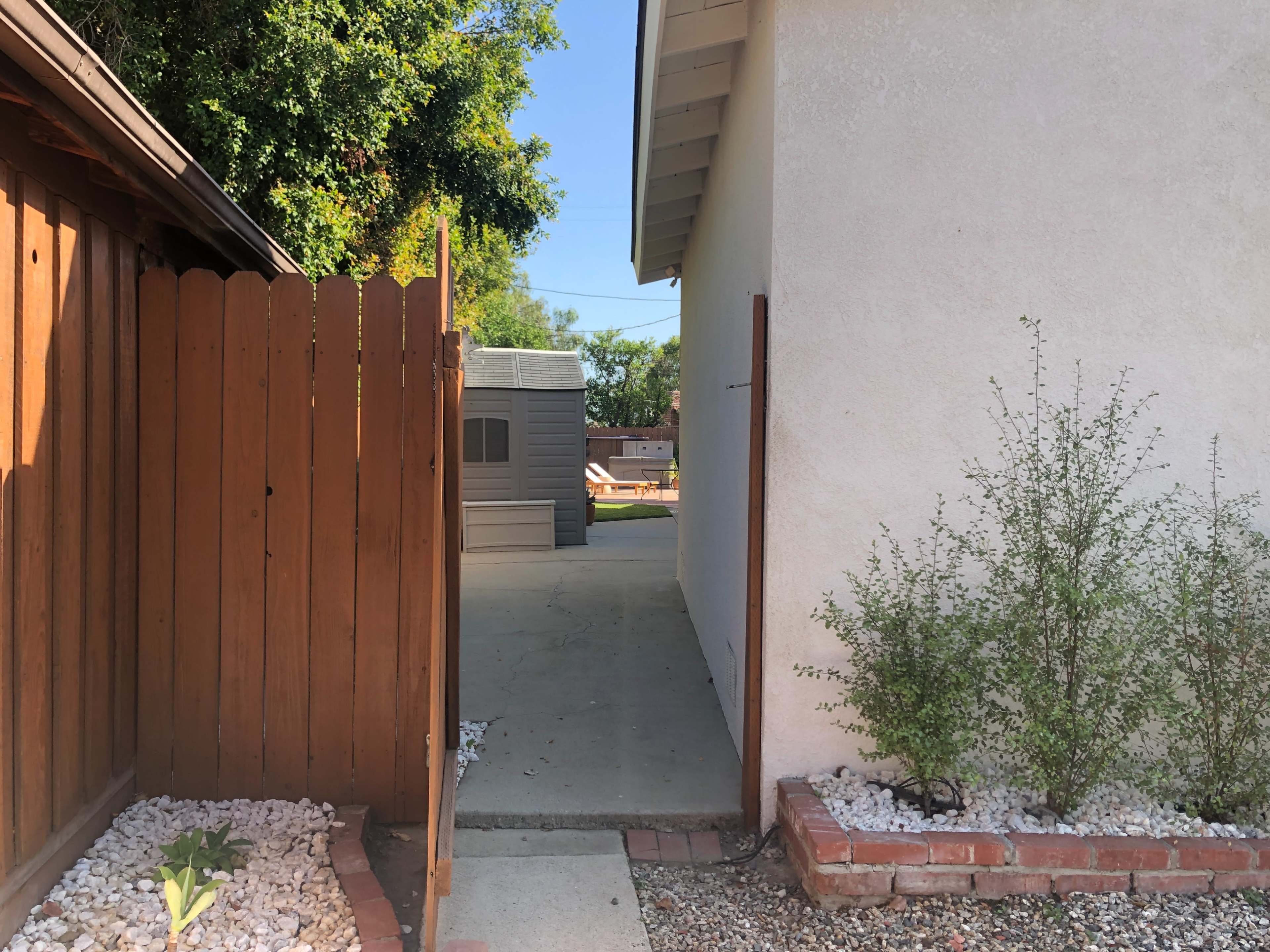 A narrow pathway leads from a wooden fence to a concrete area beside a house, flanked by decorative stones and low shrubs.