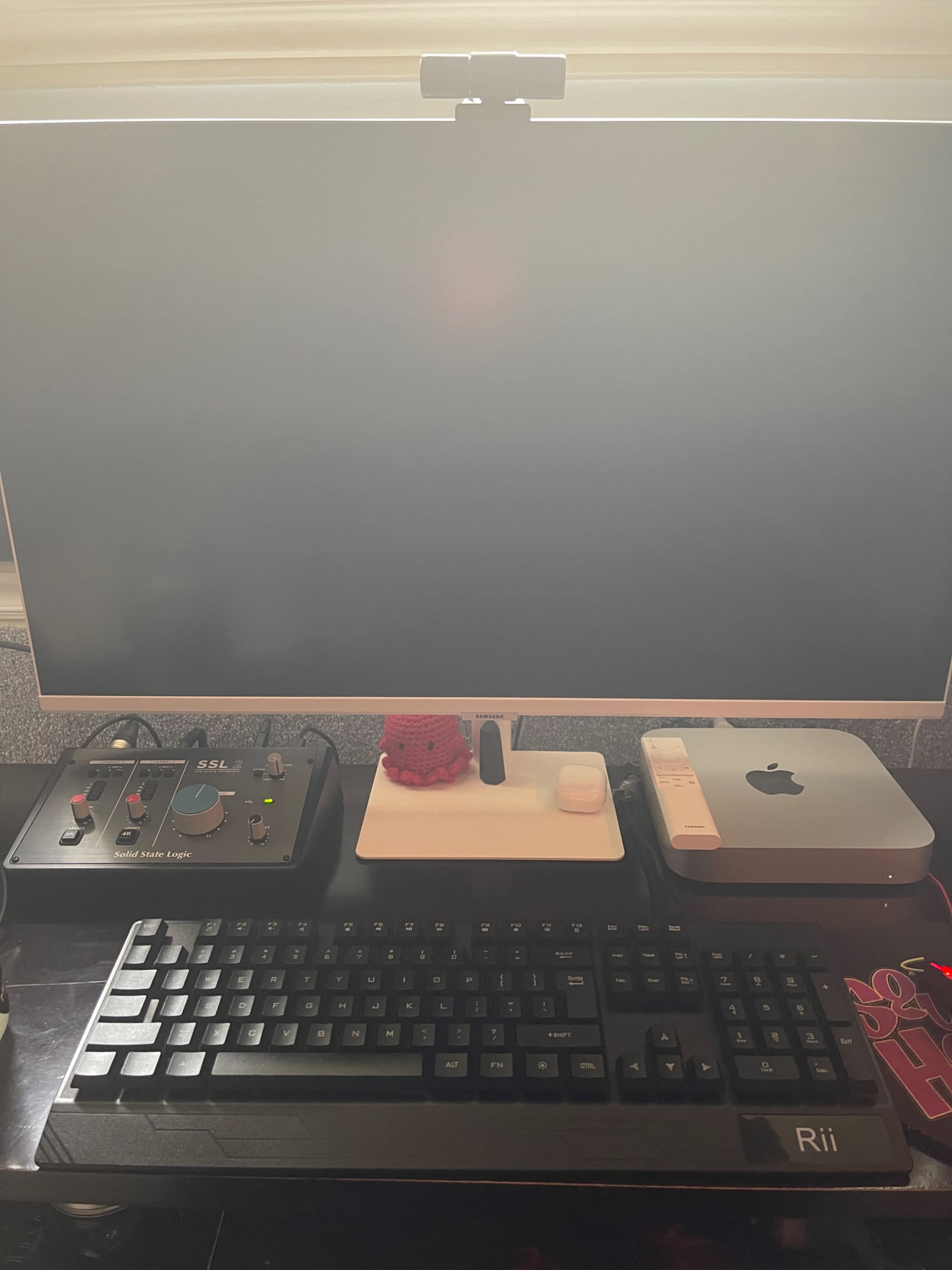 A black keyboard sits in front of a large monitor, accompanied by a microphone, a small white device, and an Apple computer on a desk.