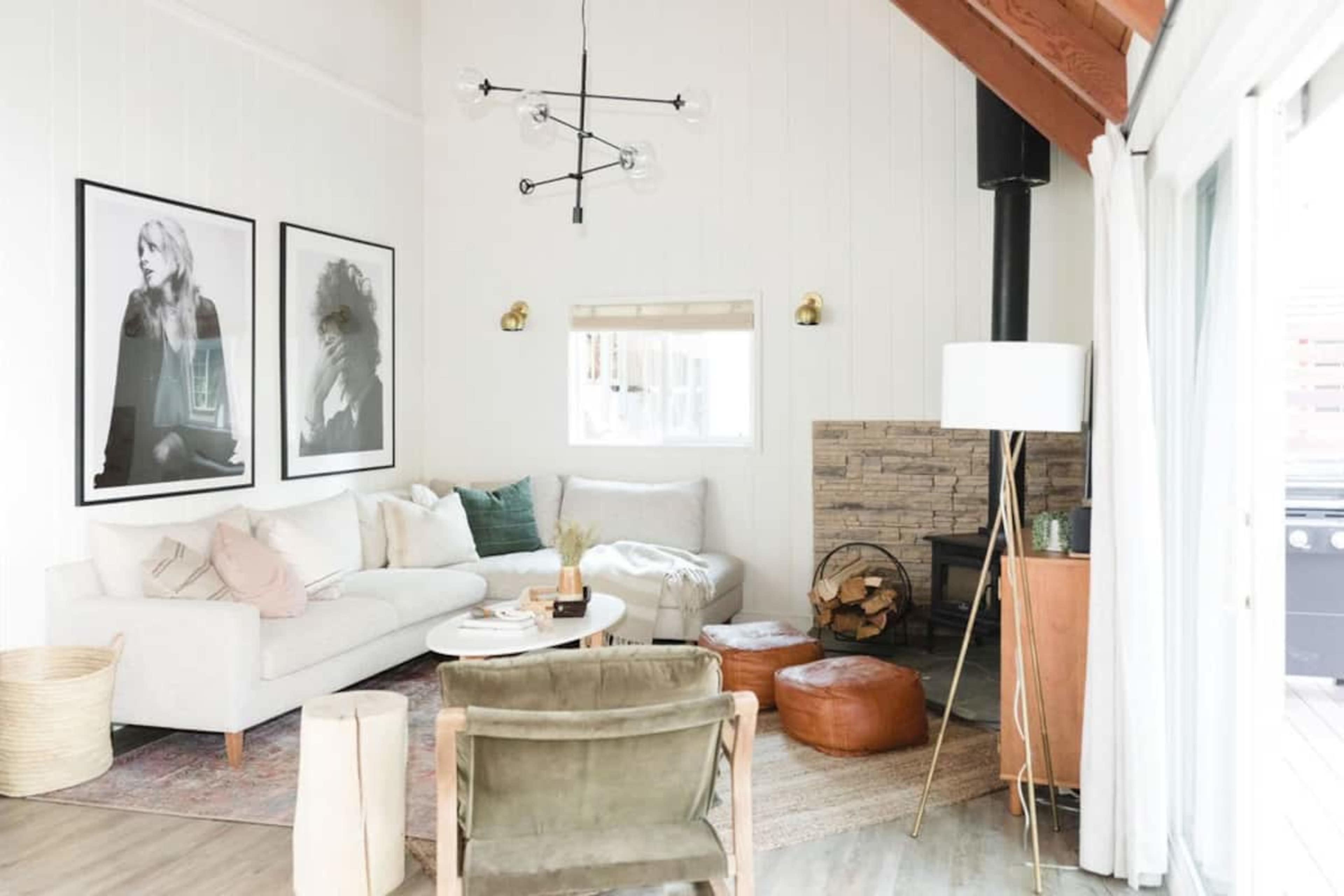 A light-filled living room features a white sofa, round coffee table, and a fireplace surrounded by wood, complemented by modern artwork on the walls.