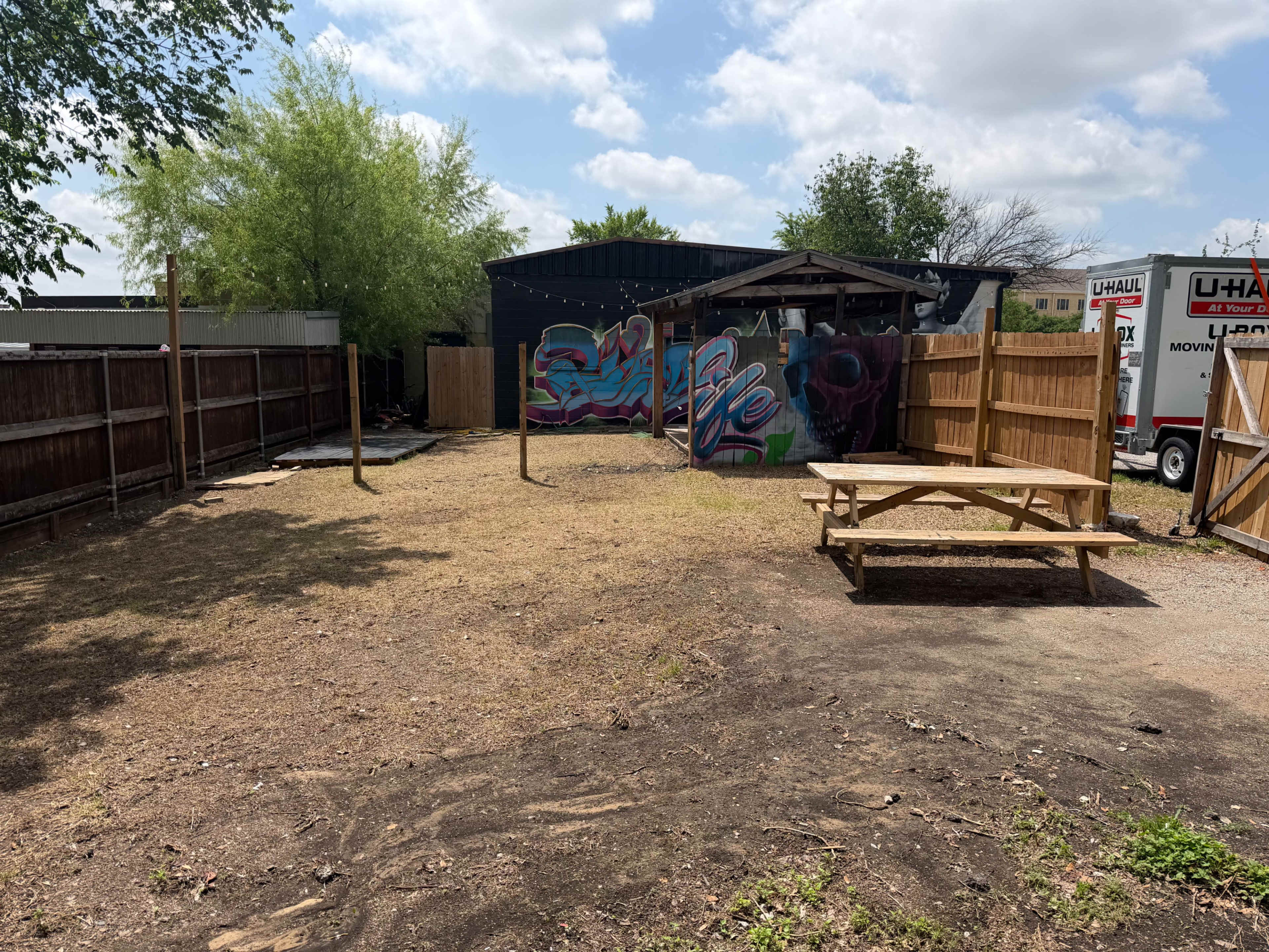 The image shows an empty outdoor space with a picnic table, surrounded by wooden fences and a mural on the back wall.