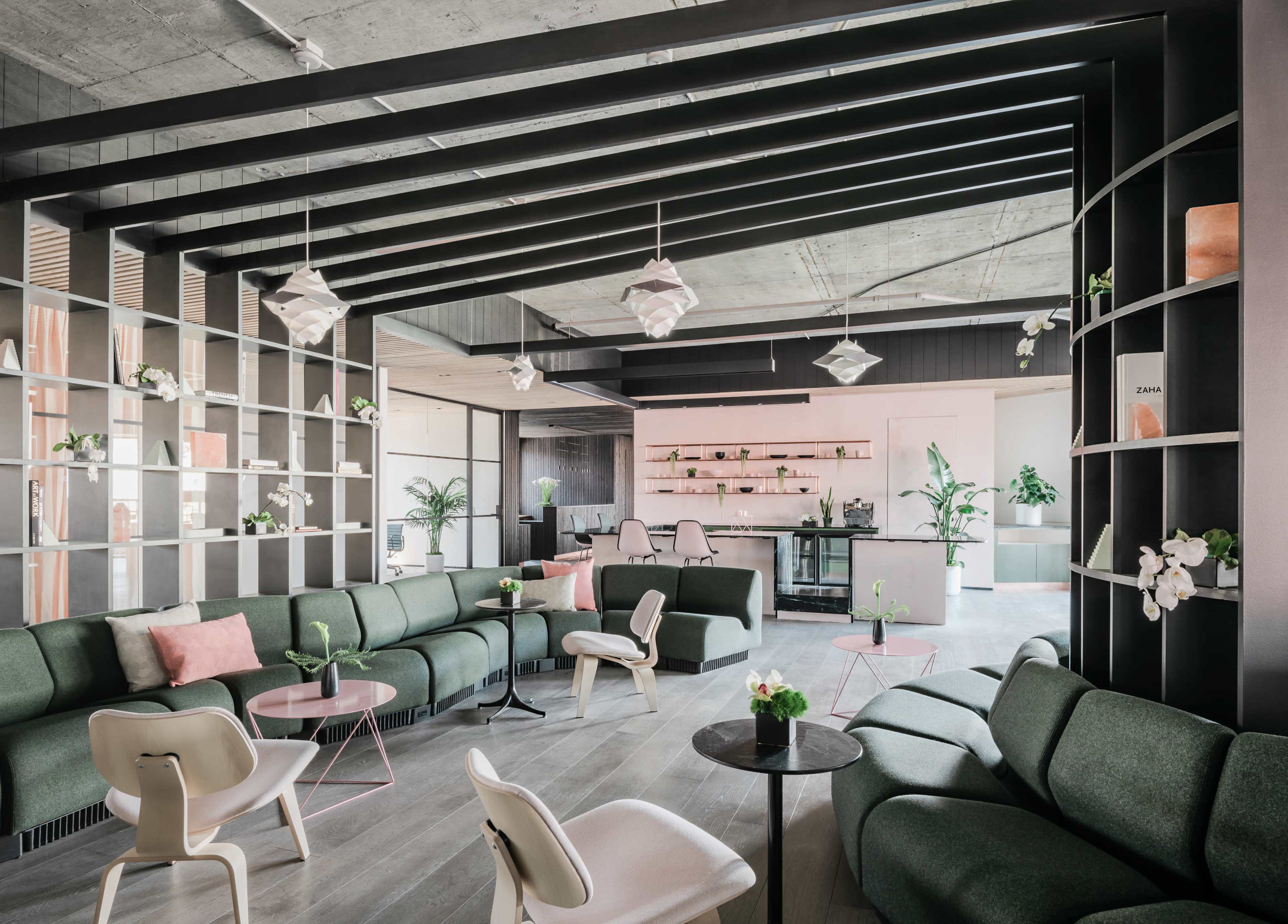 The image shows a modern lounge area featuring green sofas, white chairs, and a shelving unit with plants, set against a backdrop of a kitchen and workspace.