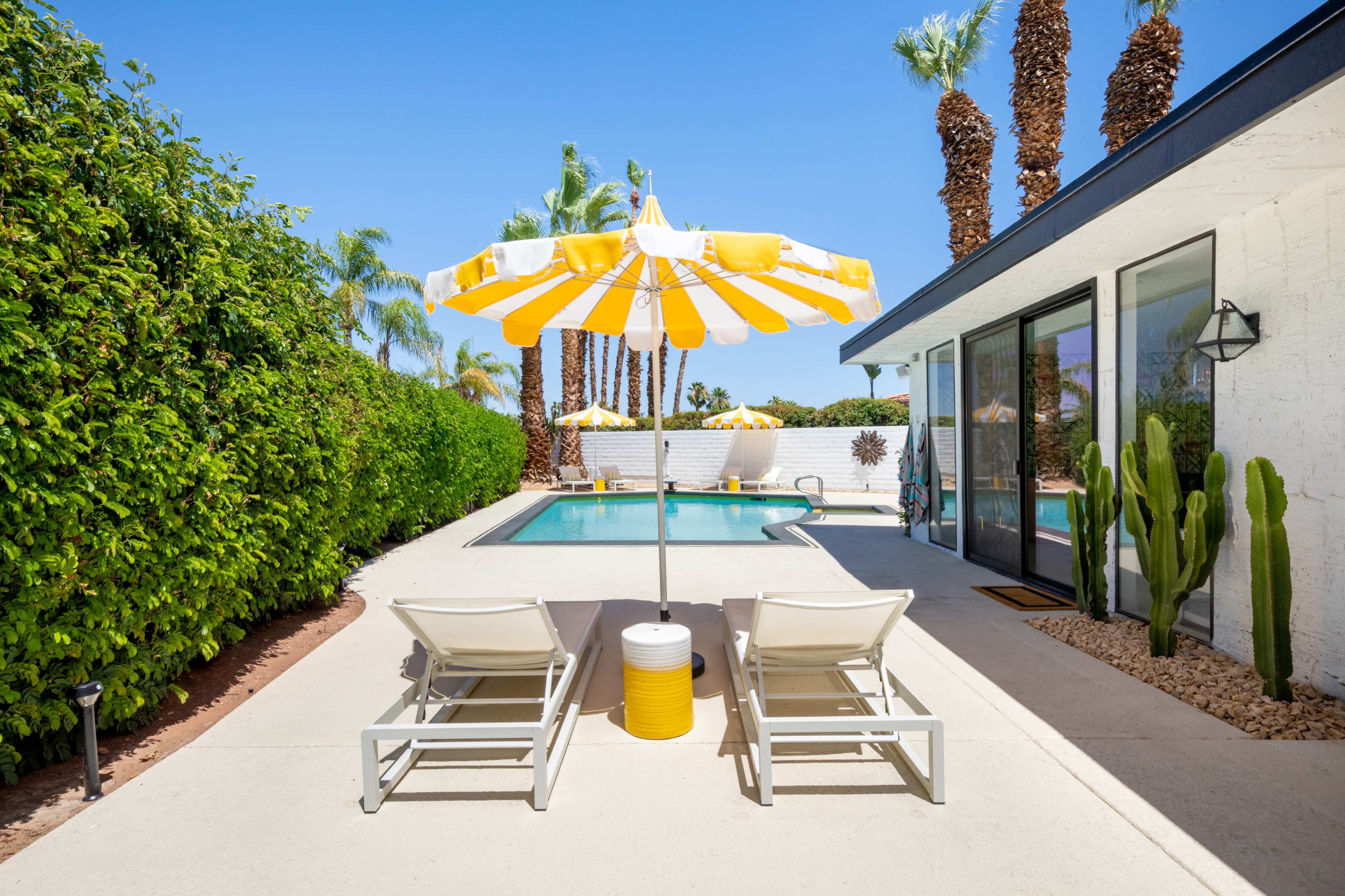 A backyard pool area features two lounge chairs beside a swimming pool, shaded by a yellow and white striped umbrella, surrounded by palm trees and a green hedge.