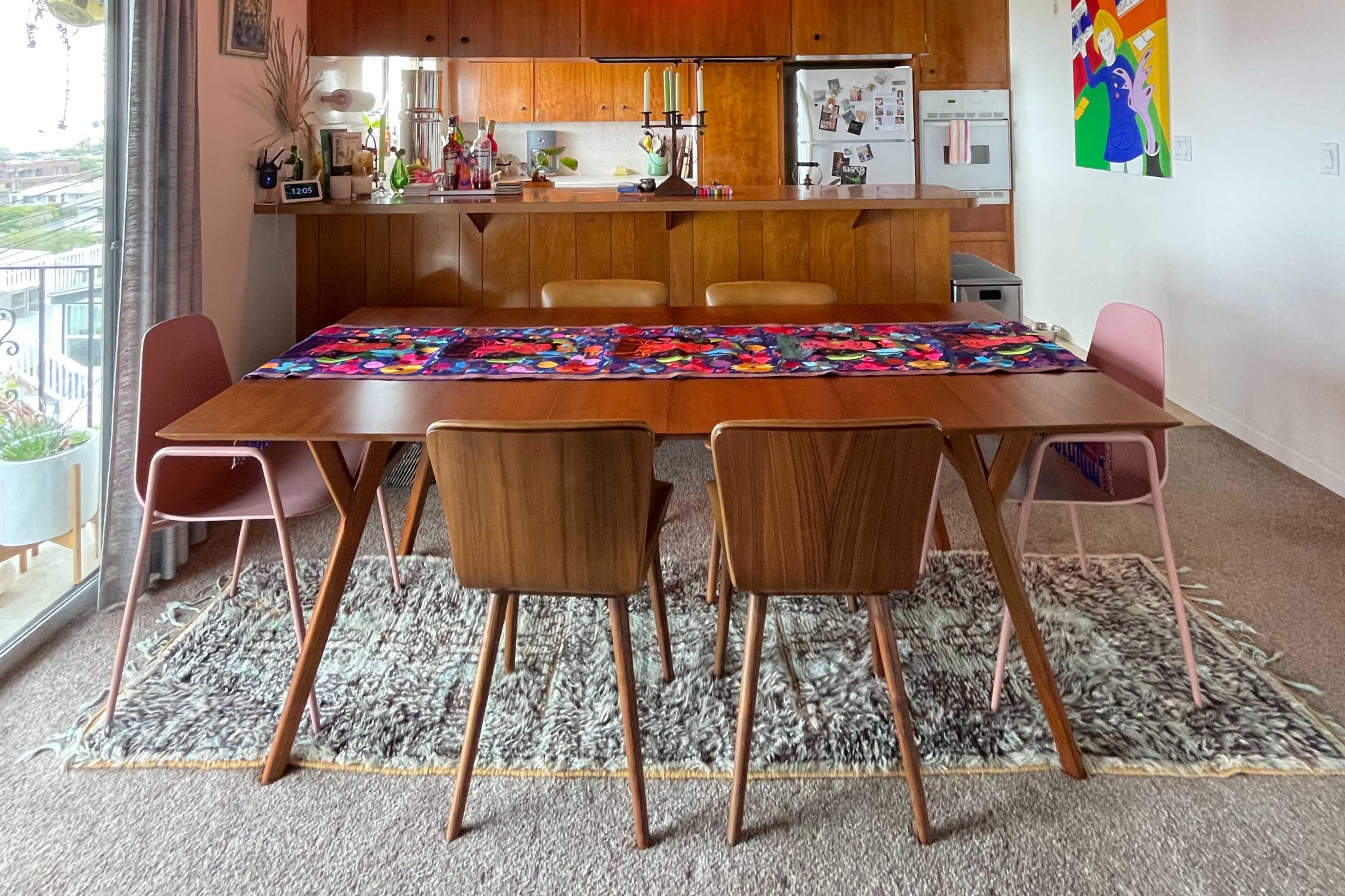 A dining table with four chairs is set in a brightly lit room featuring a kitchen area in the background.