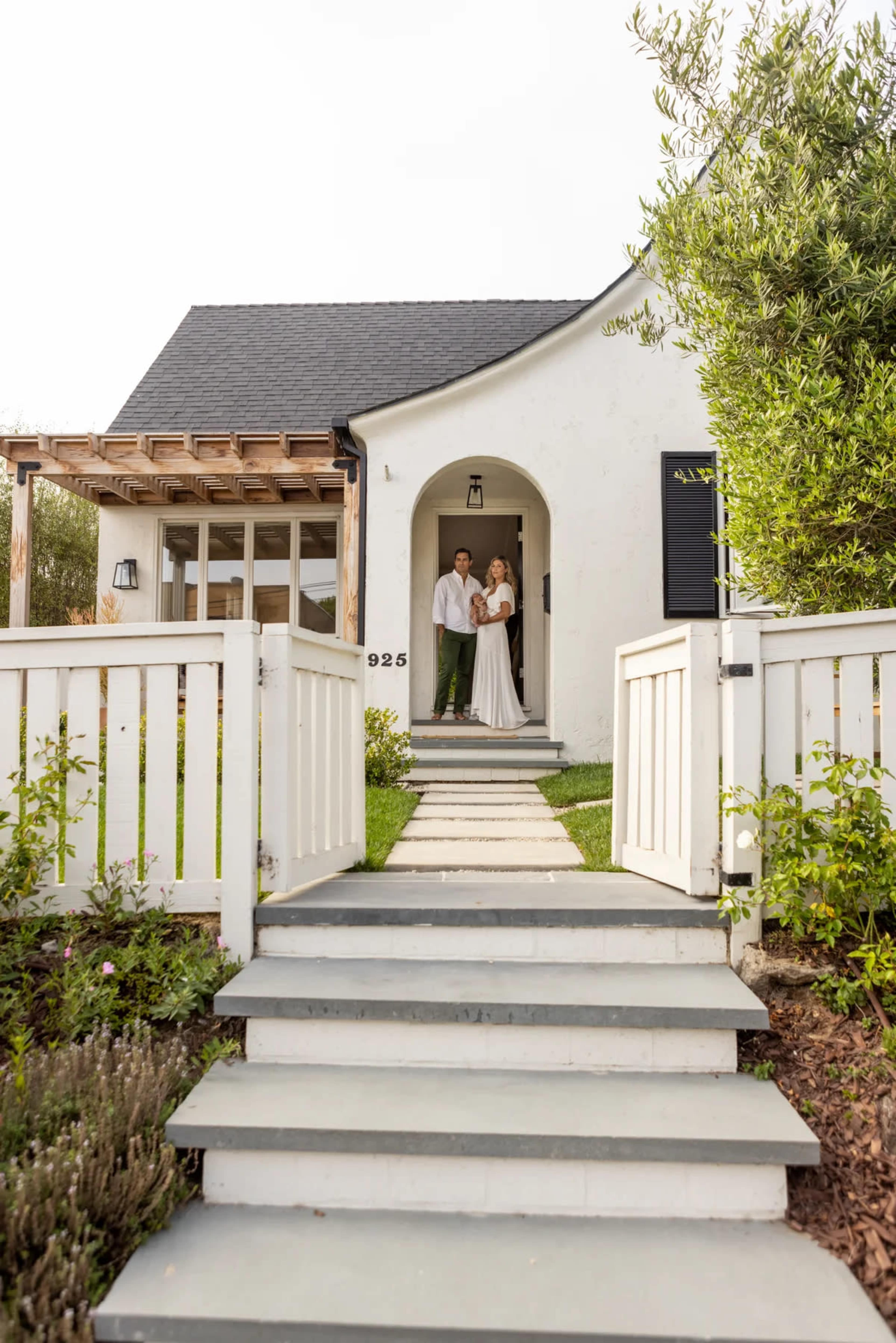 A couple stands on the steps of a white house with a black roof, framed by a white picket fence and greenery.