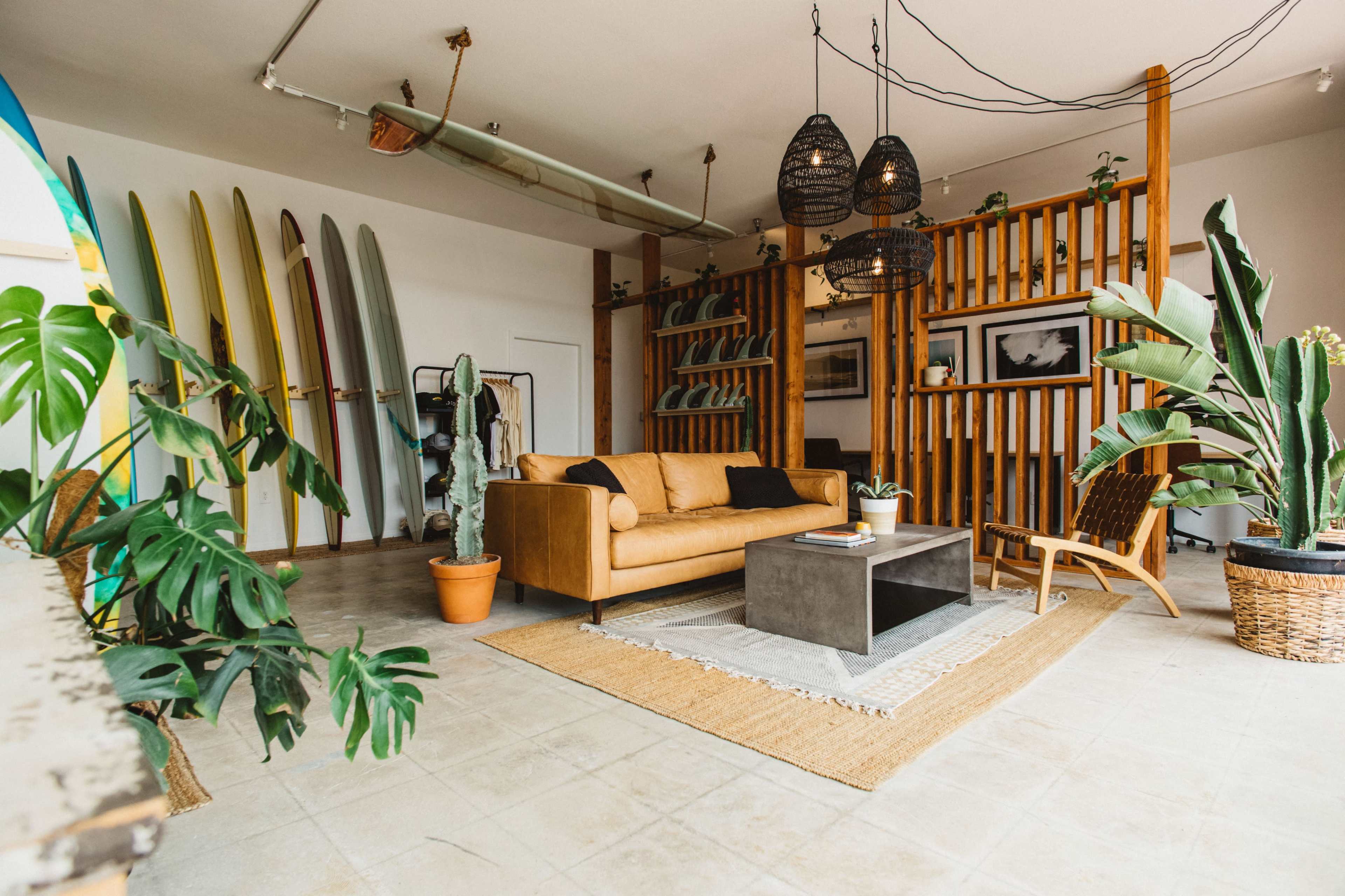 A modern lounge area features a brown sofa, a concrete coffee table, and surfboards mounted on the wall, complemented by an array of indoor plants.