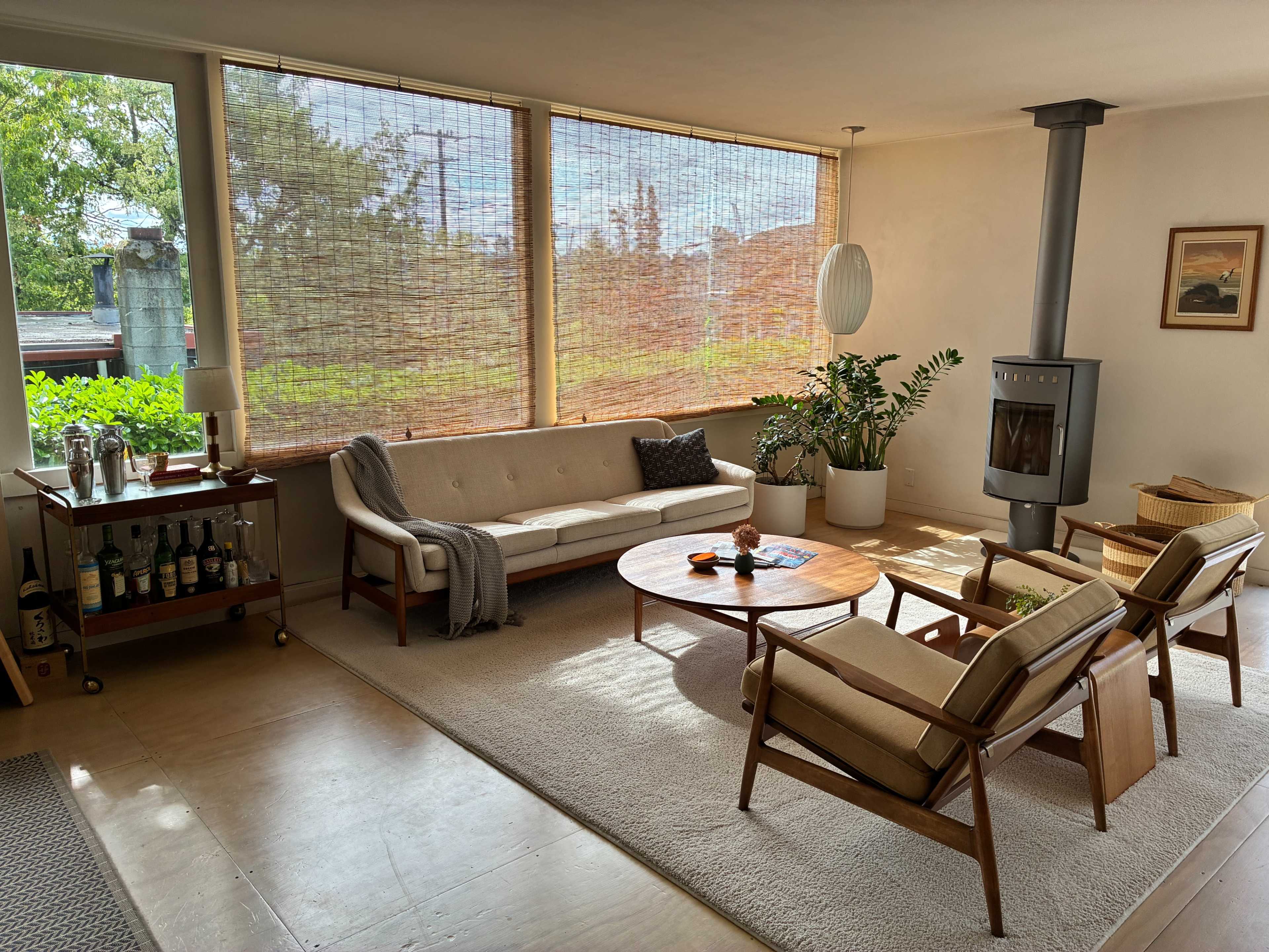 The image shows a modern living room featuring a beige sofa, two armchairs, a round coffee table, and a wood-burning stove, with large windows dressed in woven shades that allow natural light to fill the space.