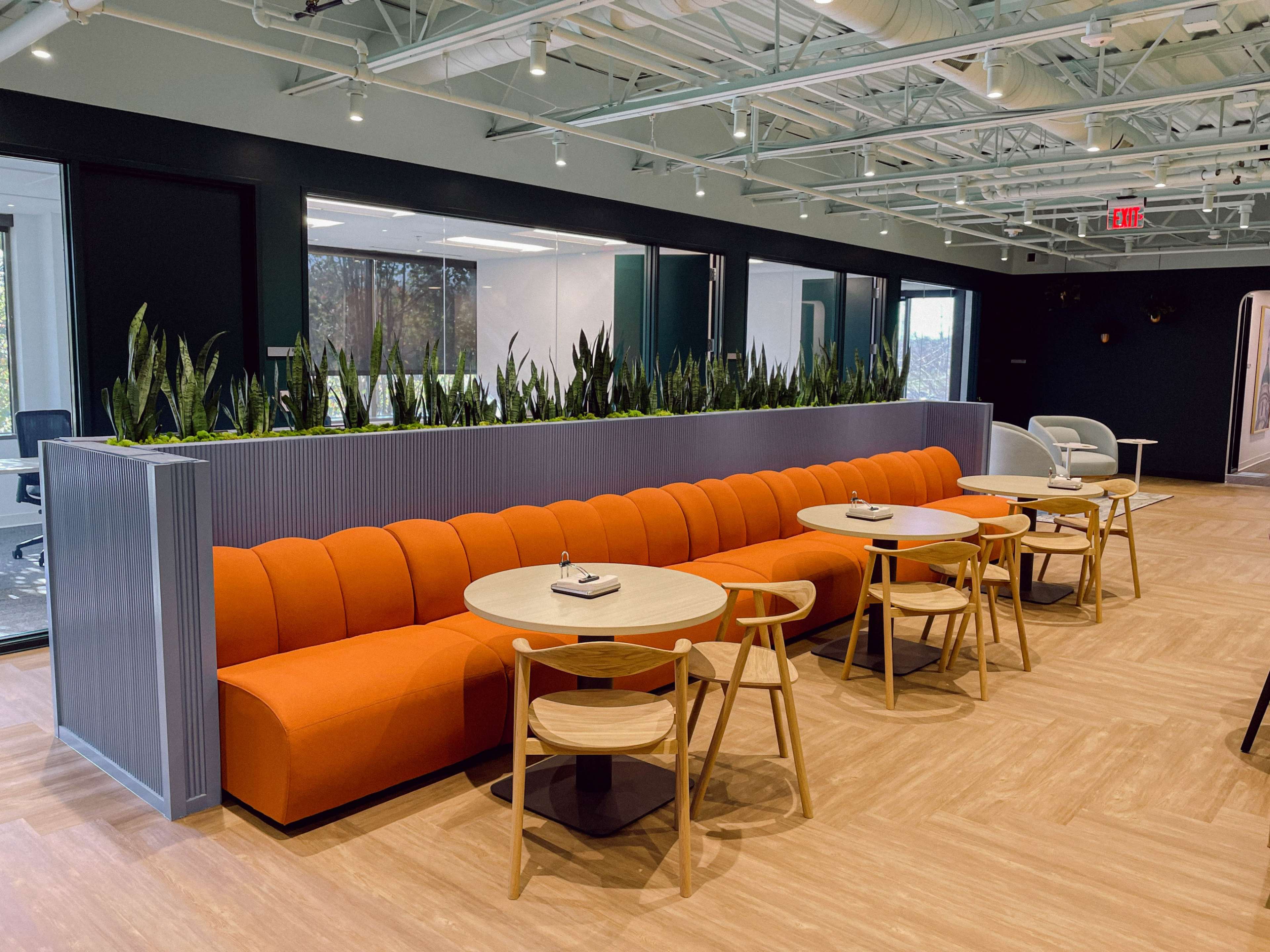 A modern interior space featuring an orange upholstered sofa, round tables, and wooden chairs arranged in a seating area surrounded by greenery.