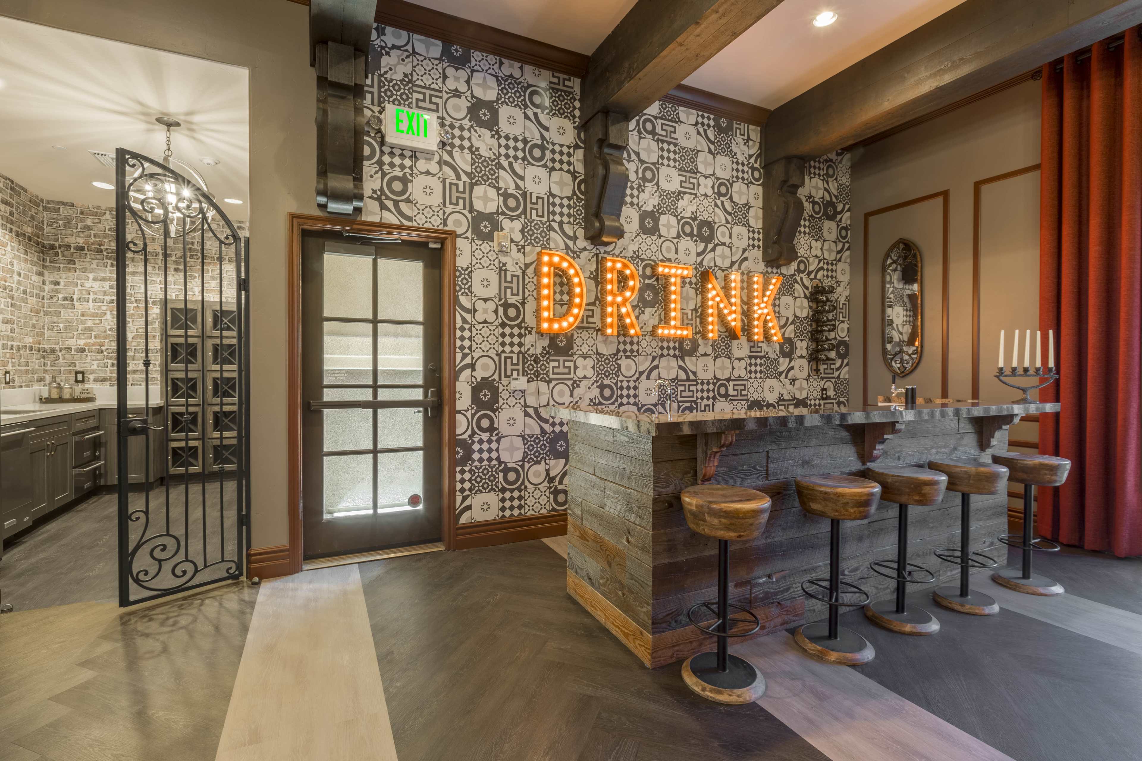 A stylish bar area with a wooden counter and stools, illuminated with the word "DRINK" made of bright lights on a patterned wall.
