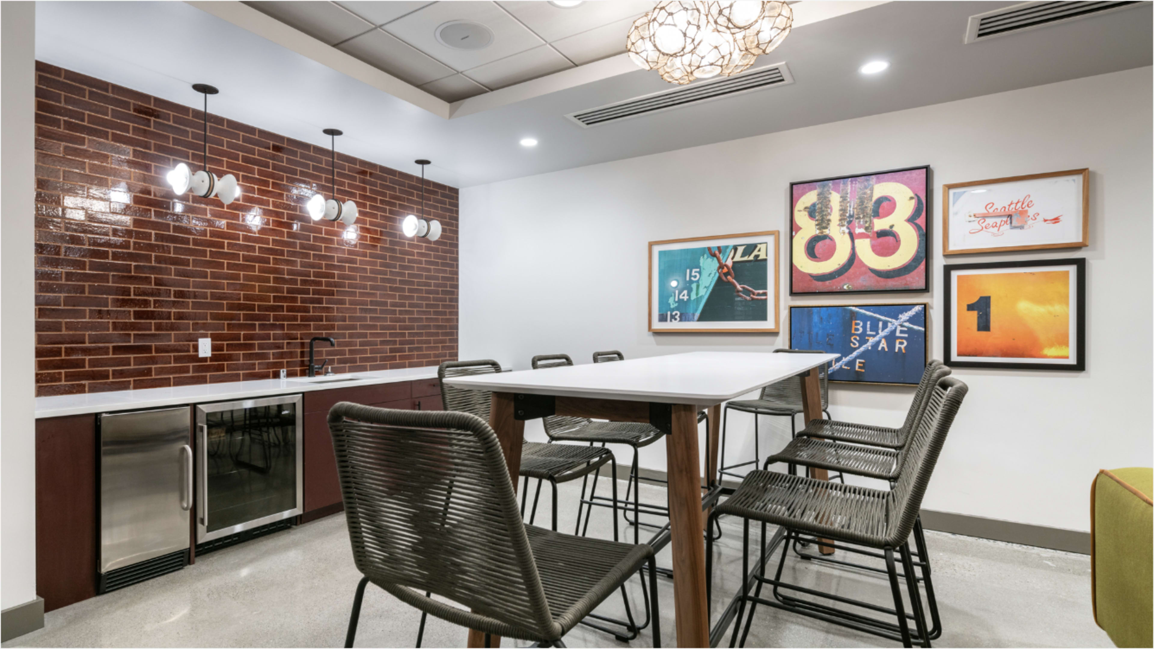 The image shows a modern dining area with a large table, high chairs, a kitchenette with a mini fridge, and decorative artwork on the walls.
