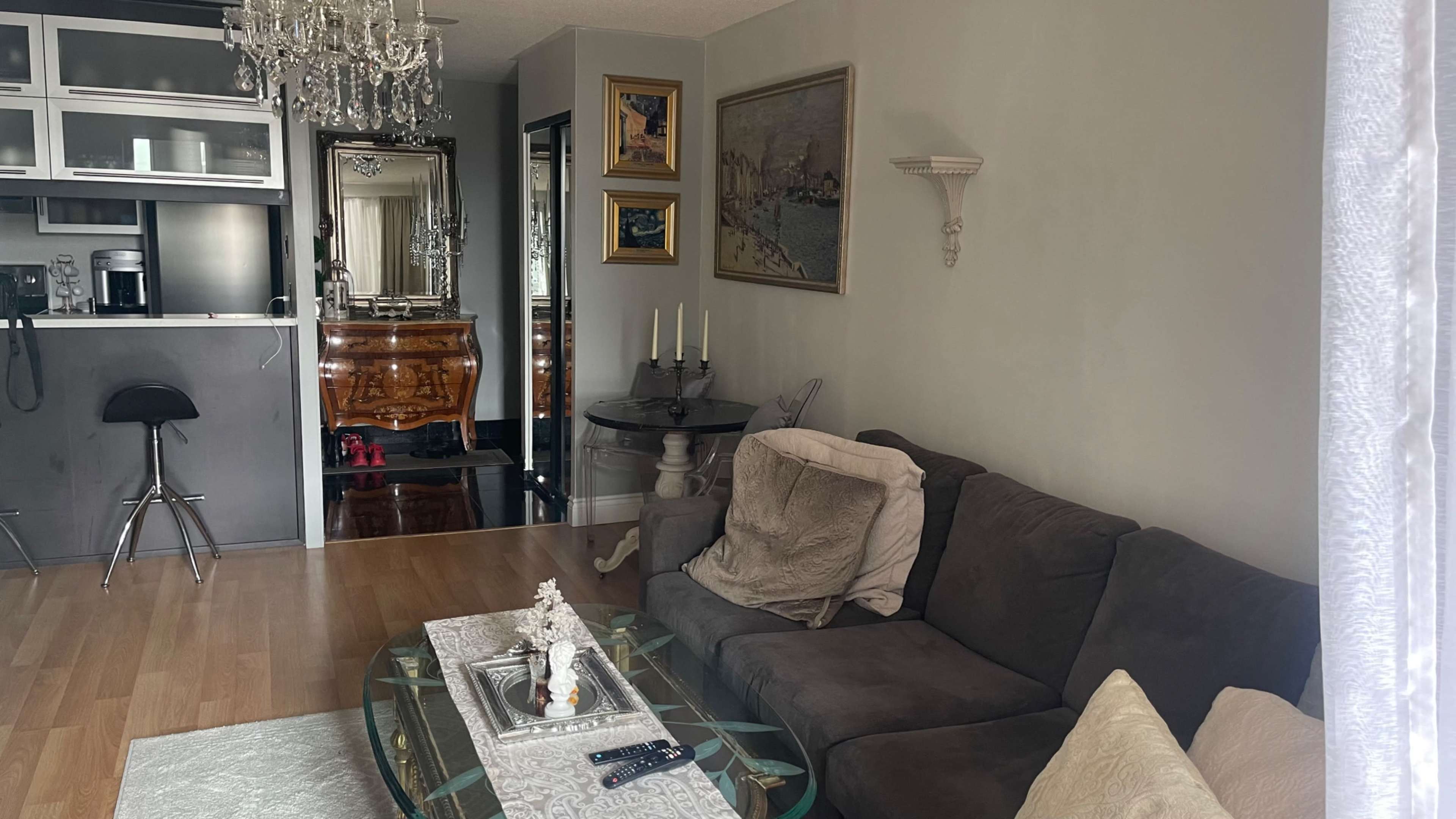 The image shows a living room with a dark sofa, a glass coffee table, decorative items, and an elegant chandelier above.