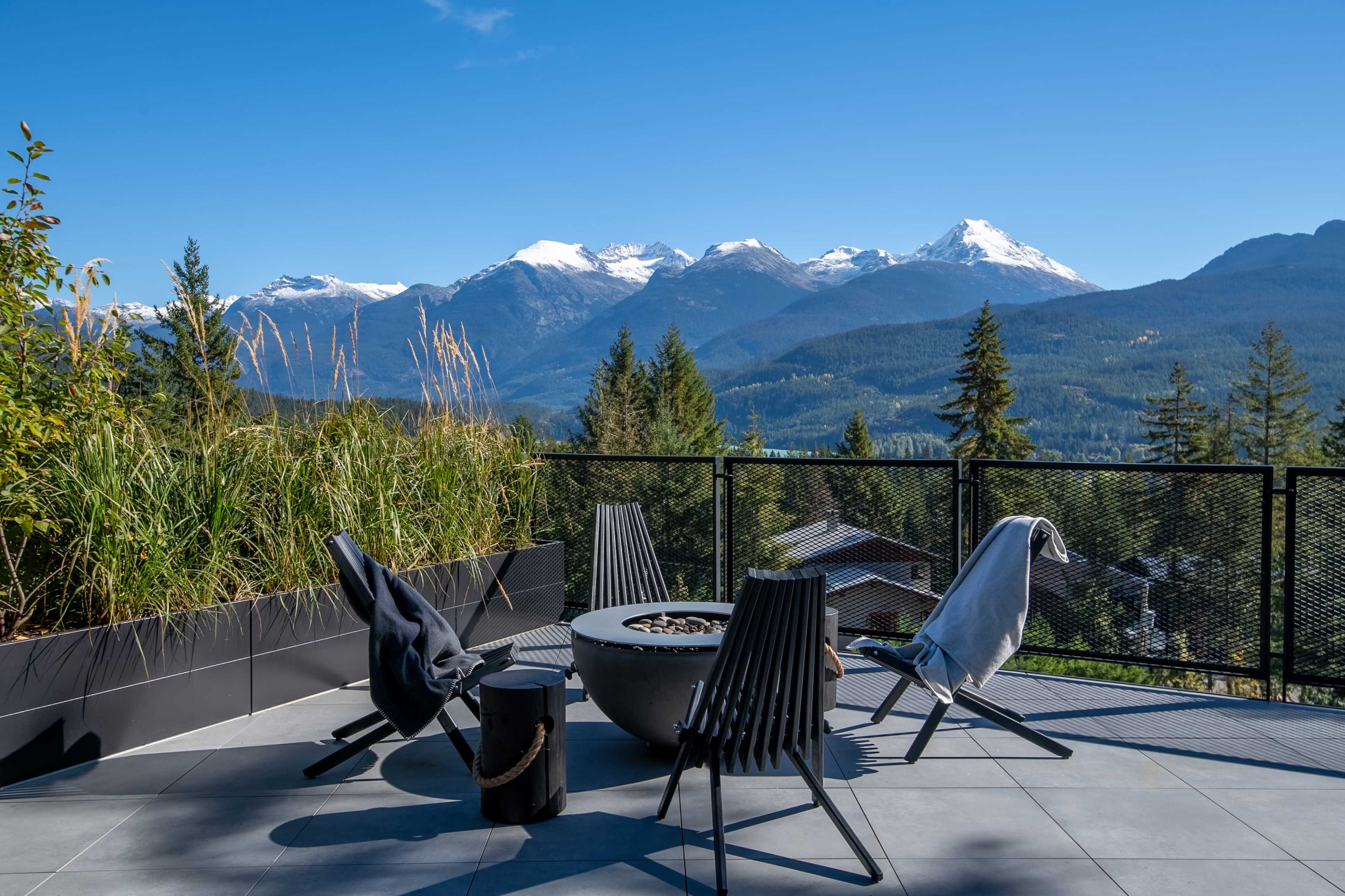 A modern outdoor patio with four chairs arranged around a circular fire pit, overlooking a mountainous landscape with snow-capped peaks.