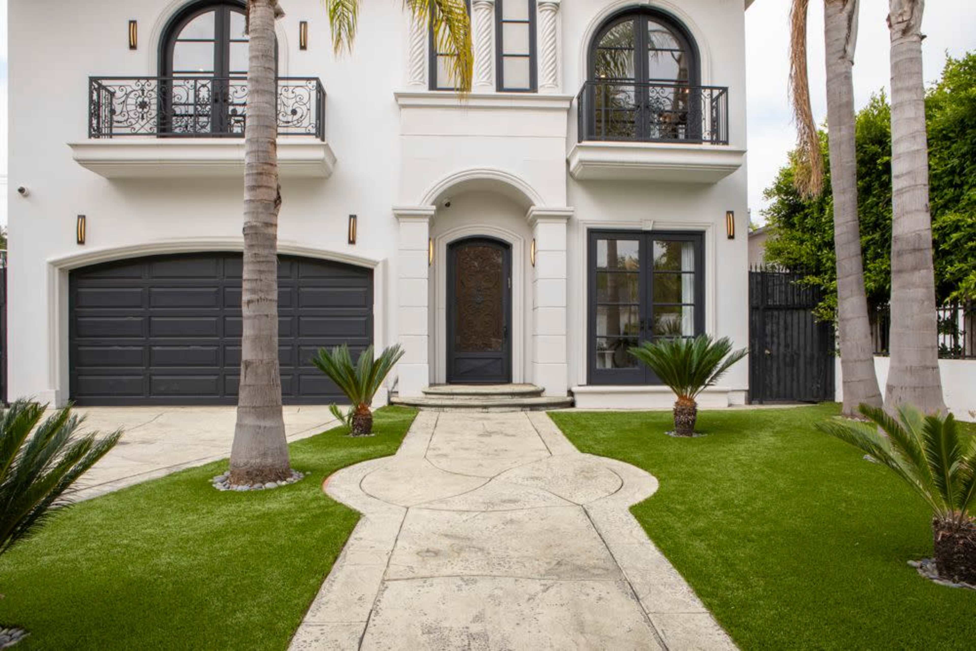 A modern white house with a decorative front door, surrounded by palm trees and a manicured lawn, with a driveway leading to a two-car garage.