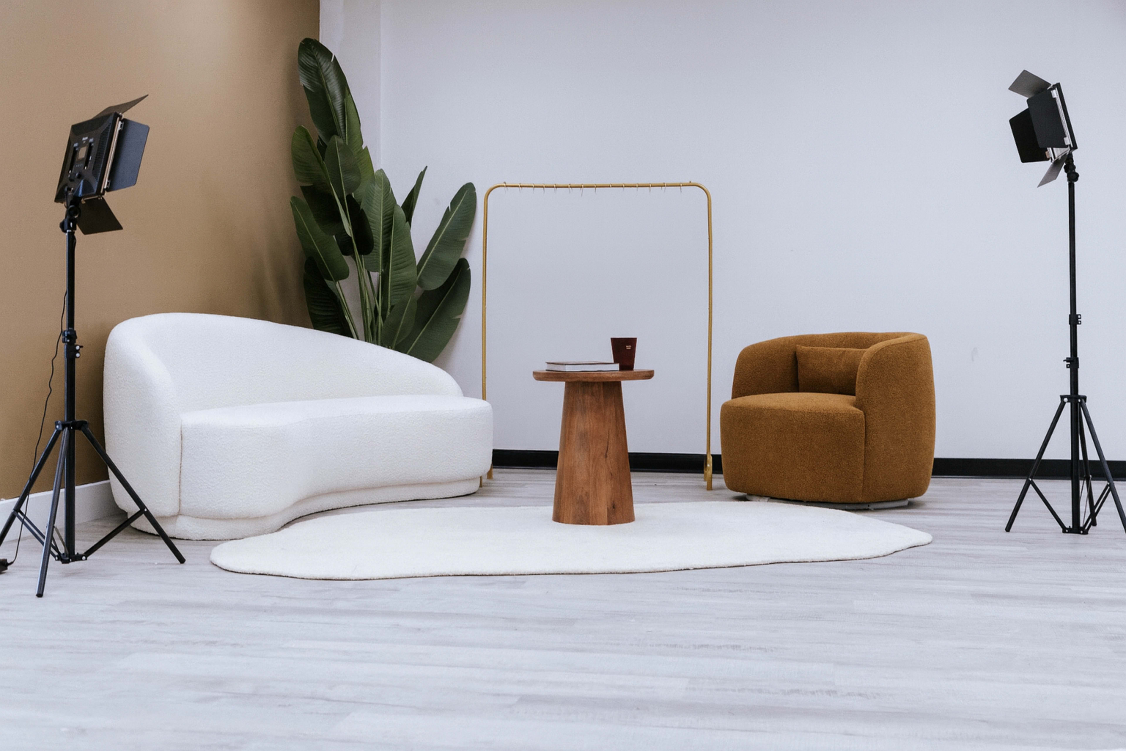 The image shows a modern, minimalist lounge setup featuring a white sofa, a brown armchair, a wooden table, and studio lighting on either side.