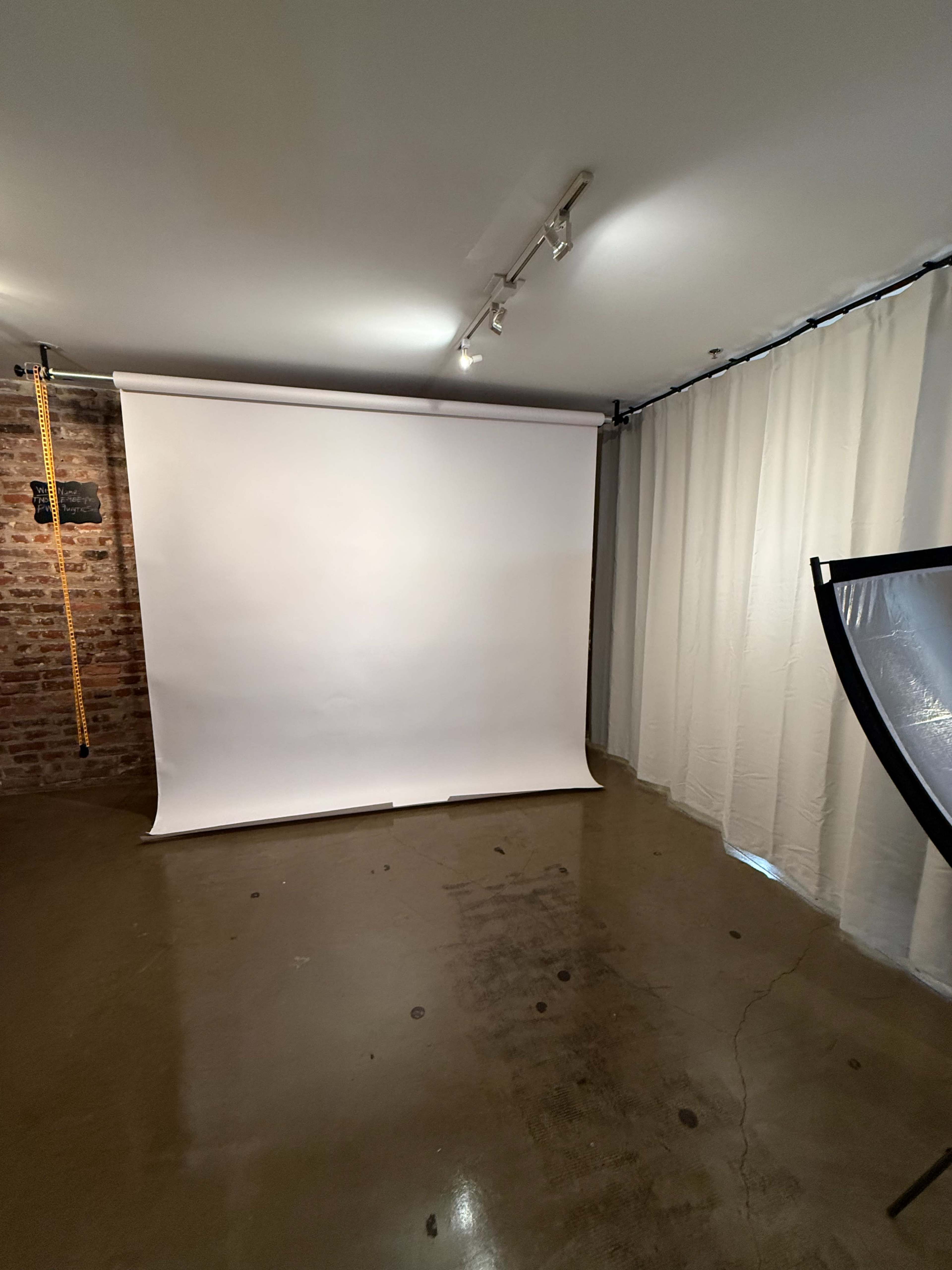 The image shows a spacious room with a plain white backdrop, brick wall, and curtain, illuminated by overhead lighting.