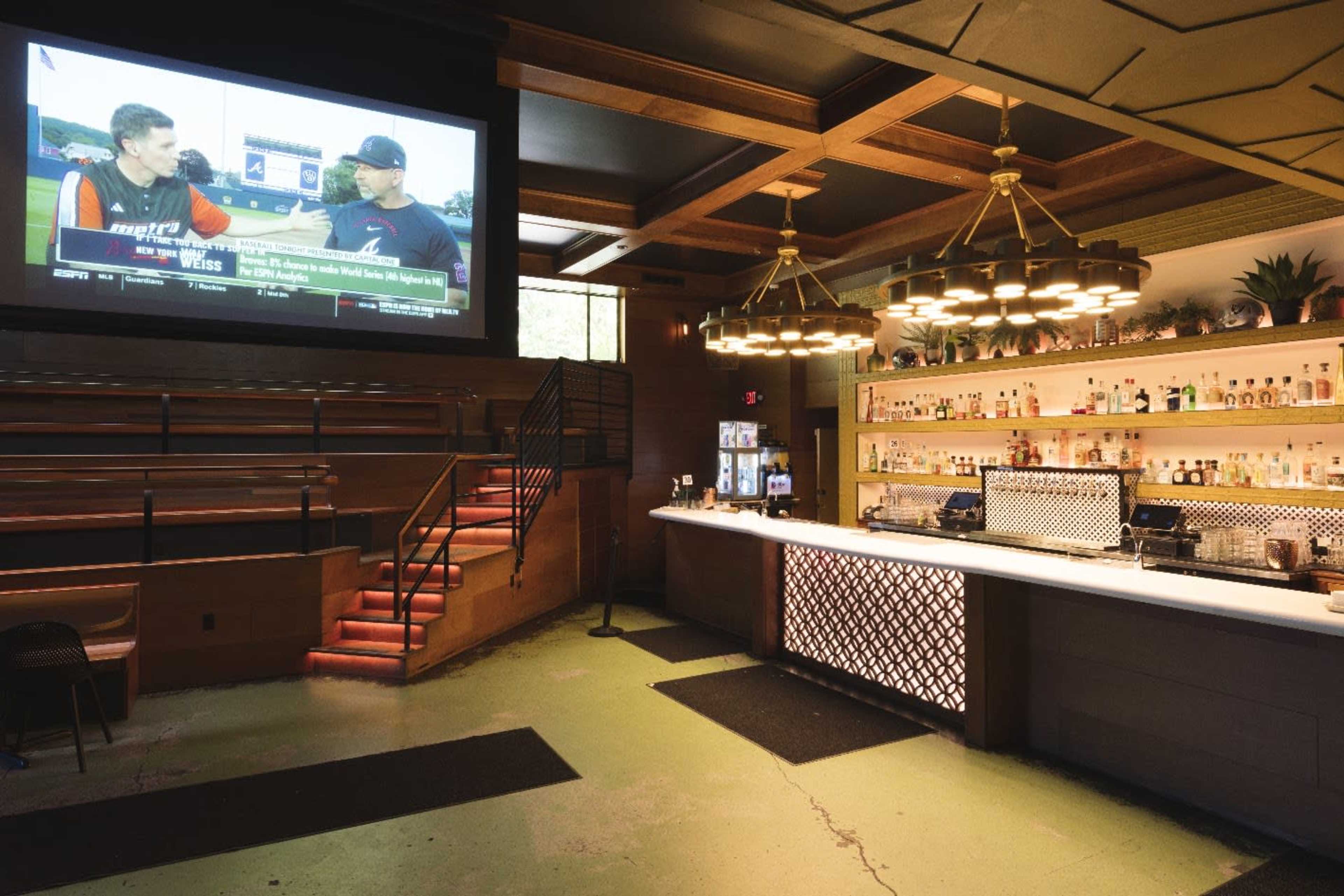 The image shows a bar area with a large screen displaying a sports program, surrounded by a well-lit counter and shelves stocked with various bottles.