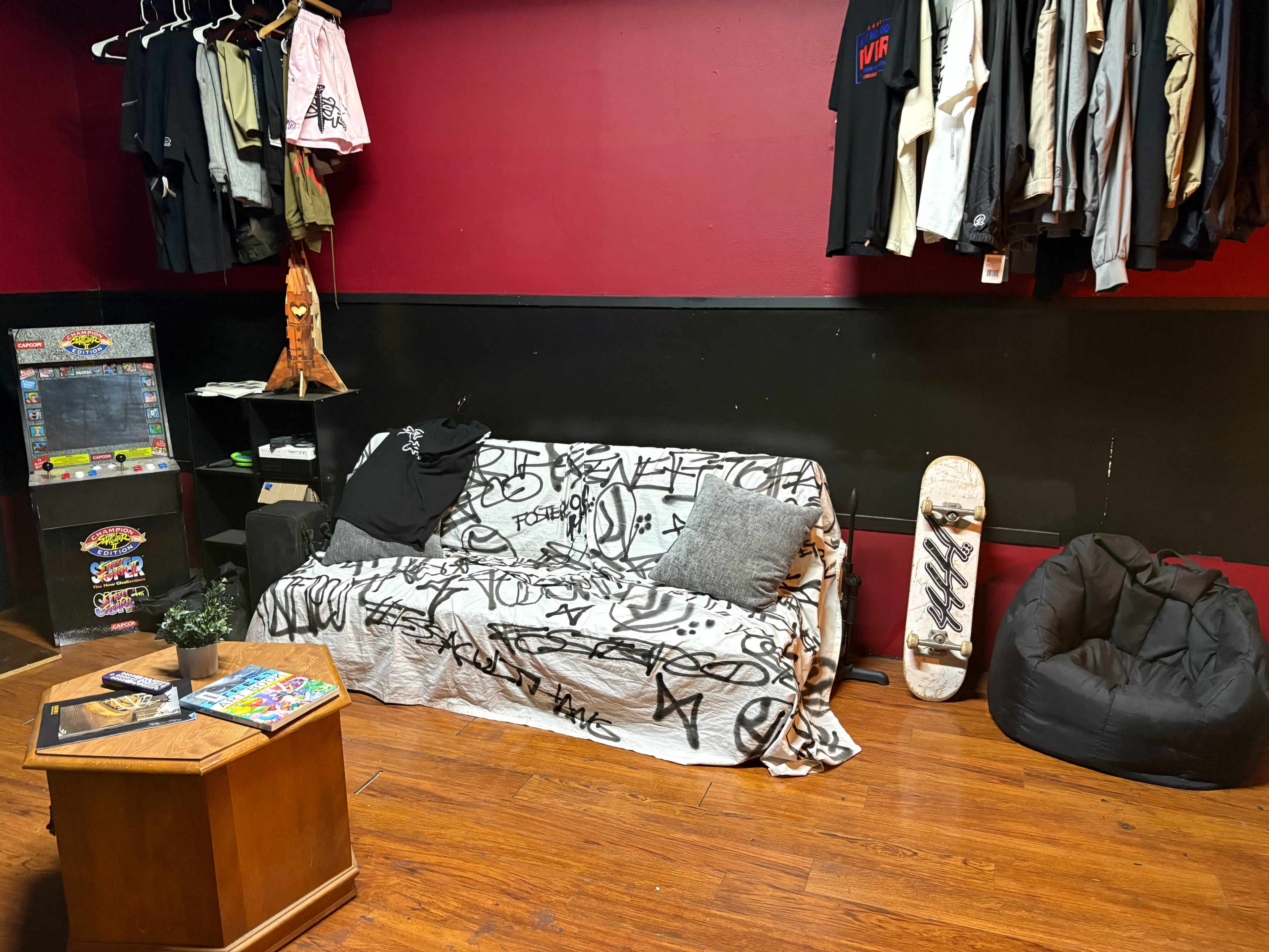 The image shows a room with a red wall, a graffiti-covered couch, a skateboard, a beanbag chair, and various clothing items hanging on a rack.