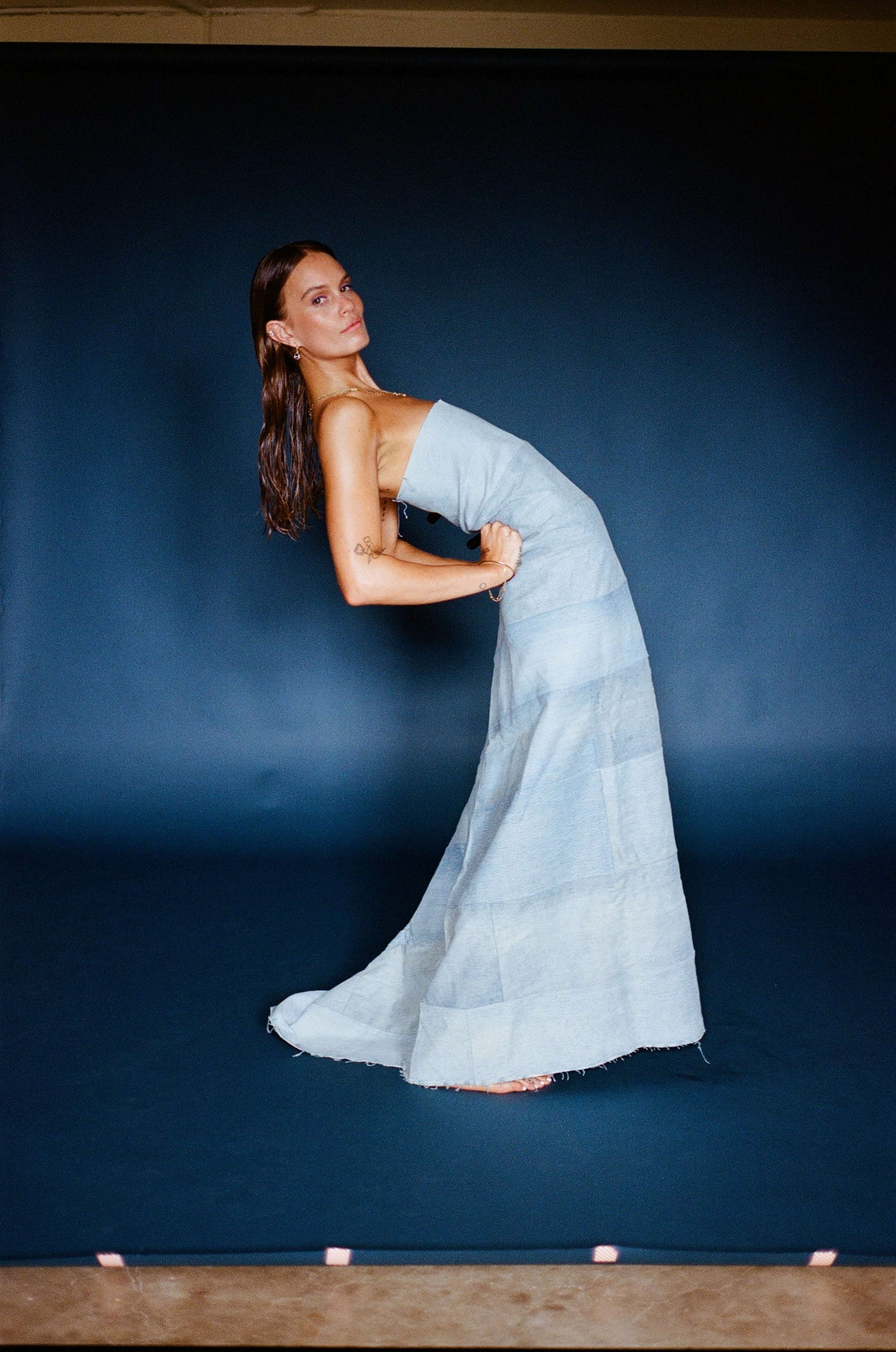 A model in a fitted, floor-length dress poses against a dark blue backdrop.