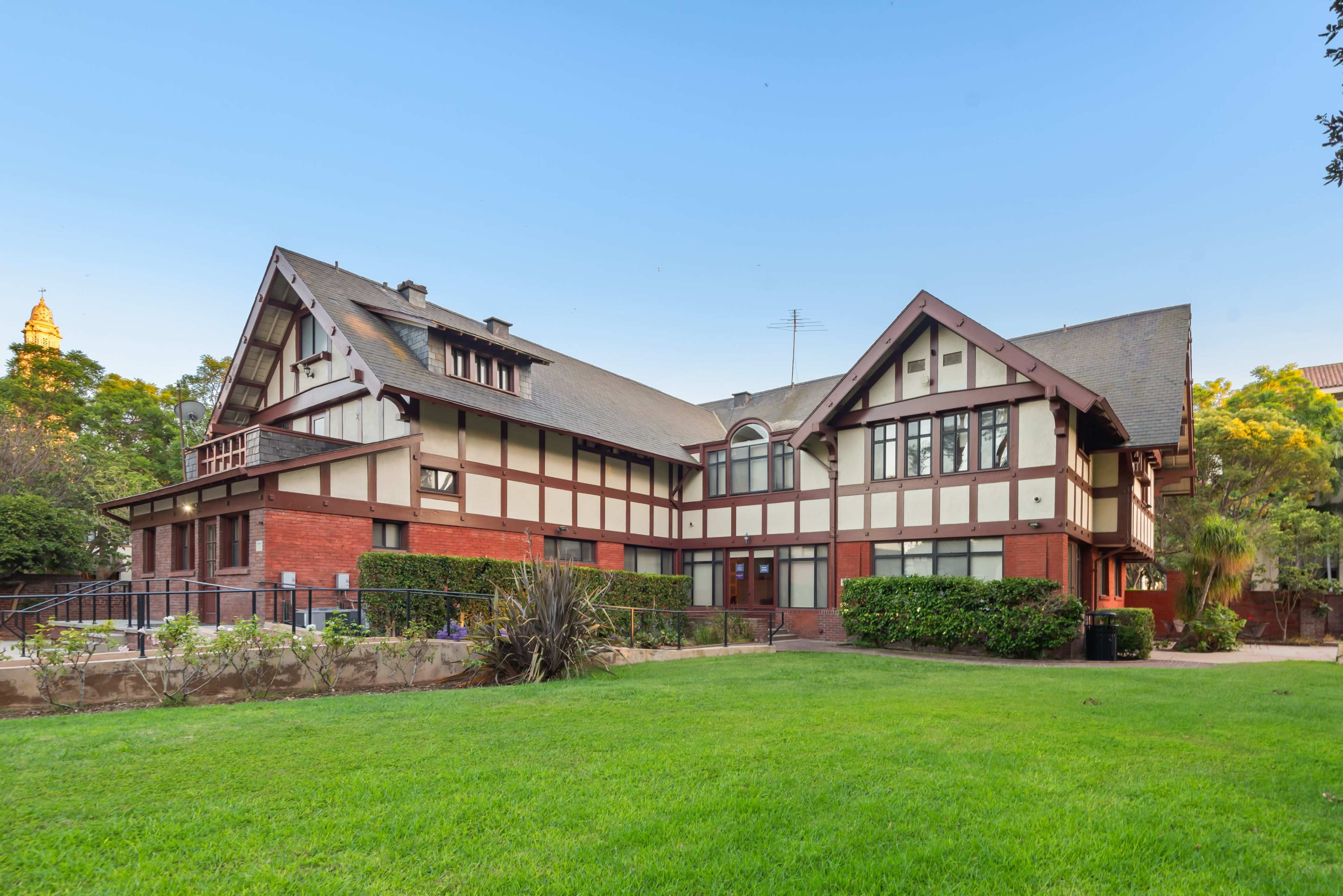The image shows a large, two-story house with a combination of brick and wood elements, situated in a grassy area with trees in the background.