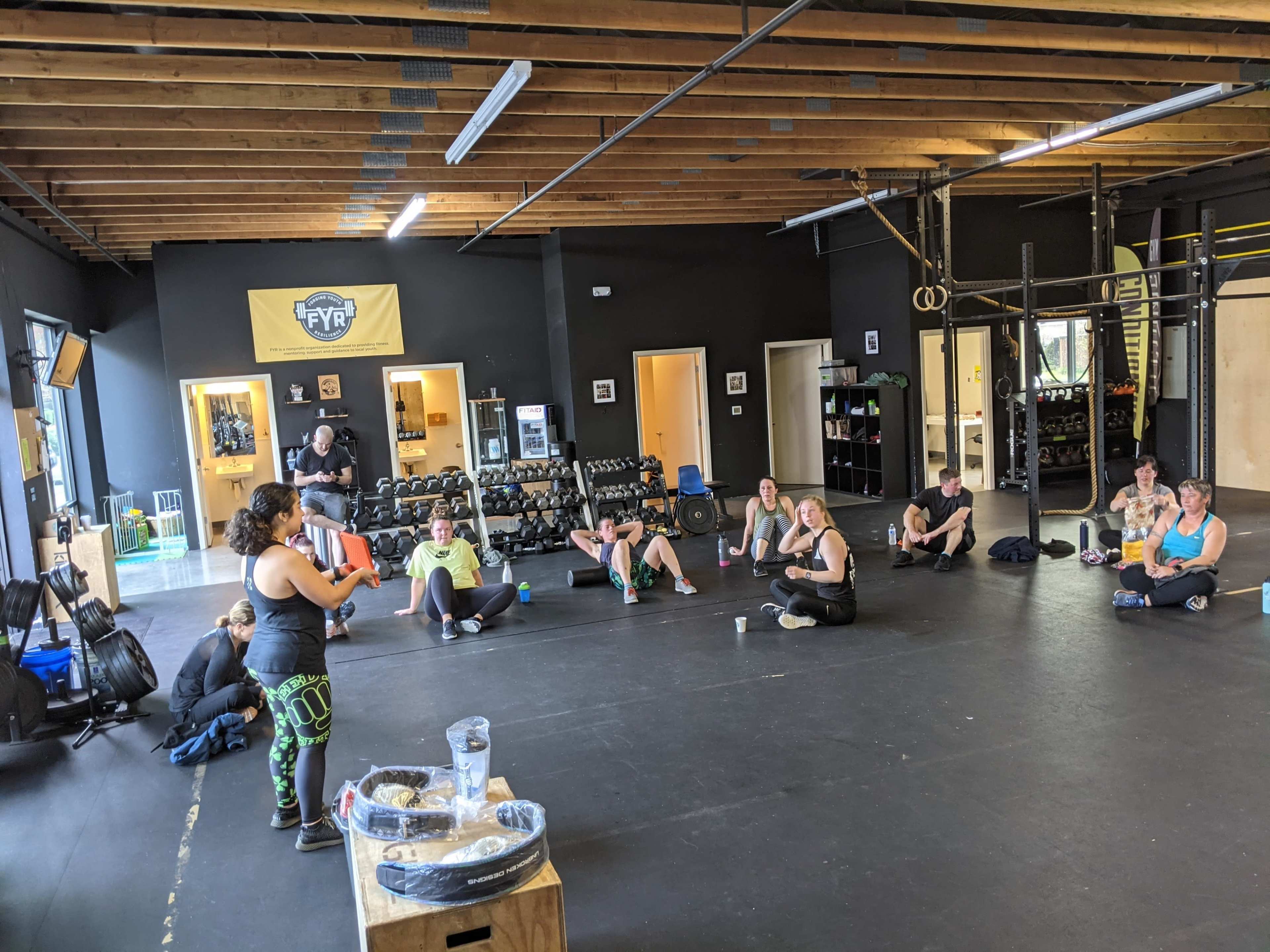 A fitness instructor demonstrates a technique to a group of participants seated on the floor in a gym.
