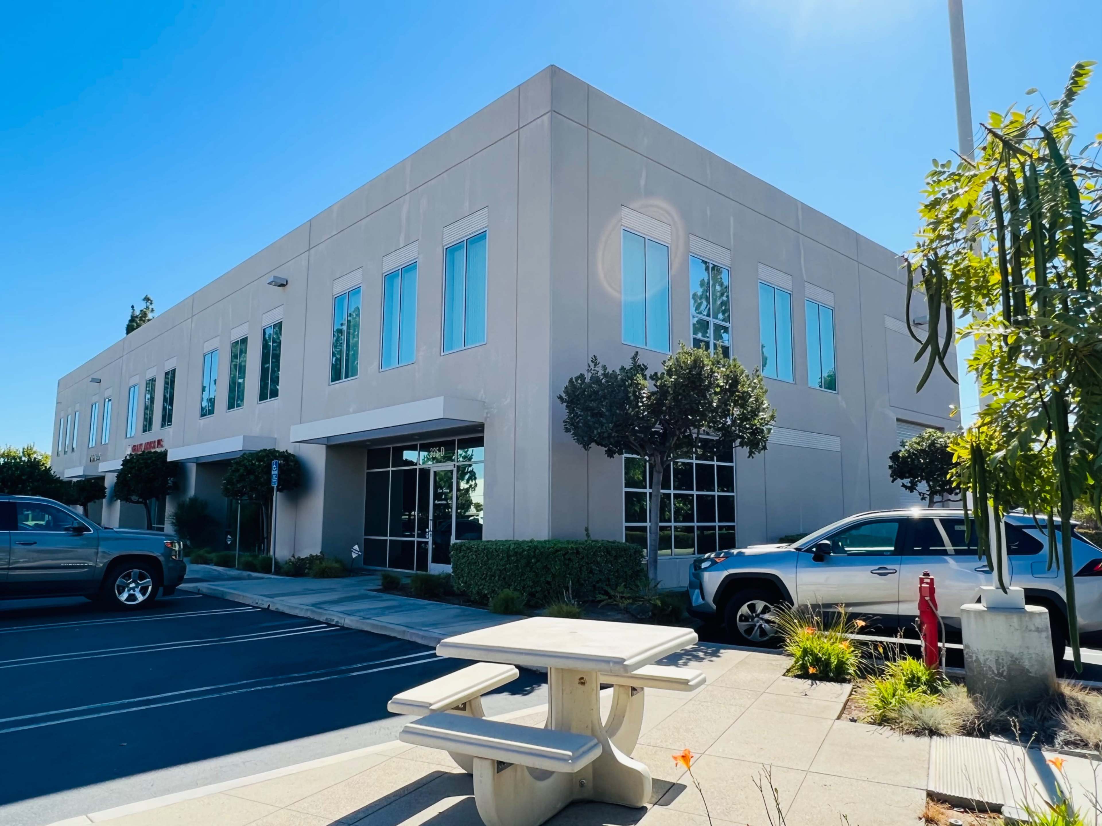 A two-story commercial building with large windows is surrounded by parked cars, landscaping, and a concrete picnic table.
