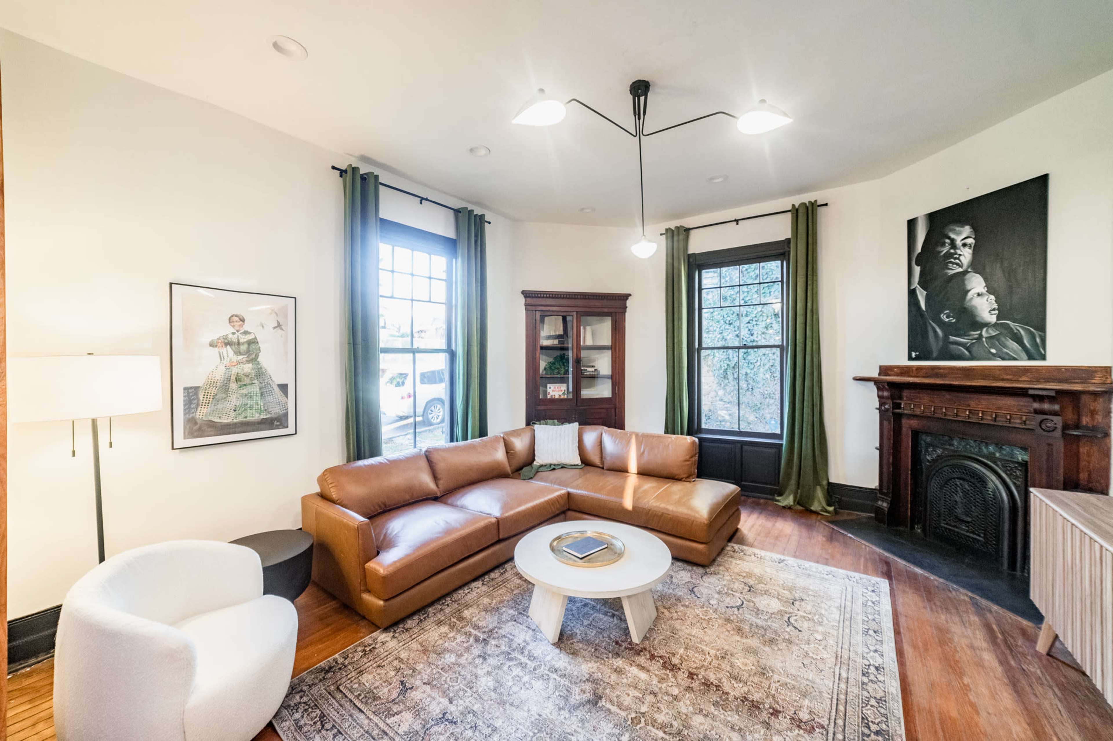 The living room features a brown leather sectional sofa, a white accent chair, and a circular coffee table on a patterned rug, with large windows and a wooden fireplace.